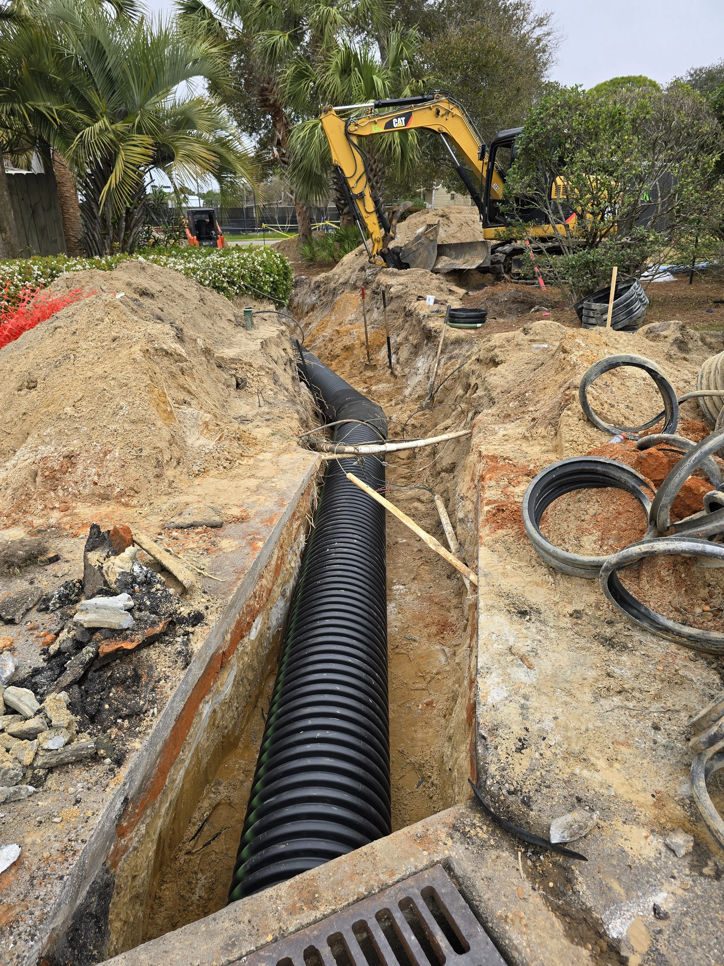 Construction site with large black drainage pipe being installed underground, surrounded by dirt and construction equipment.