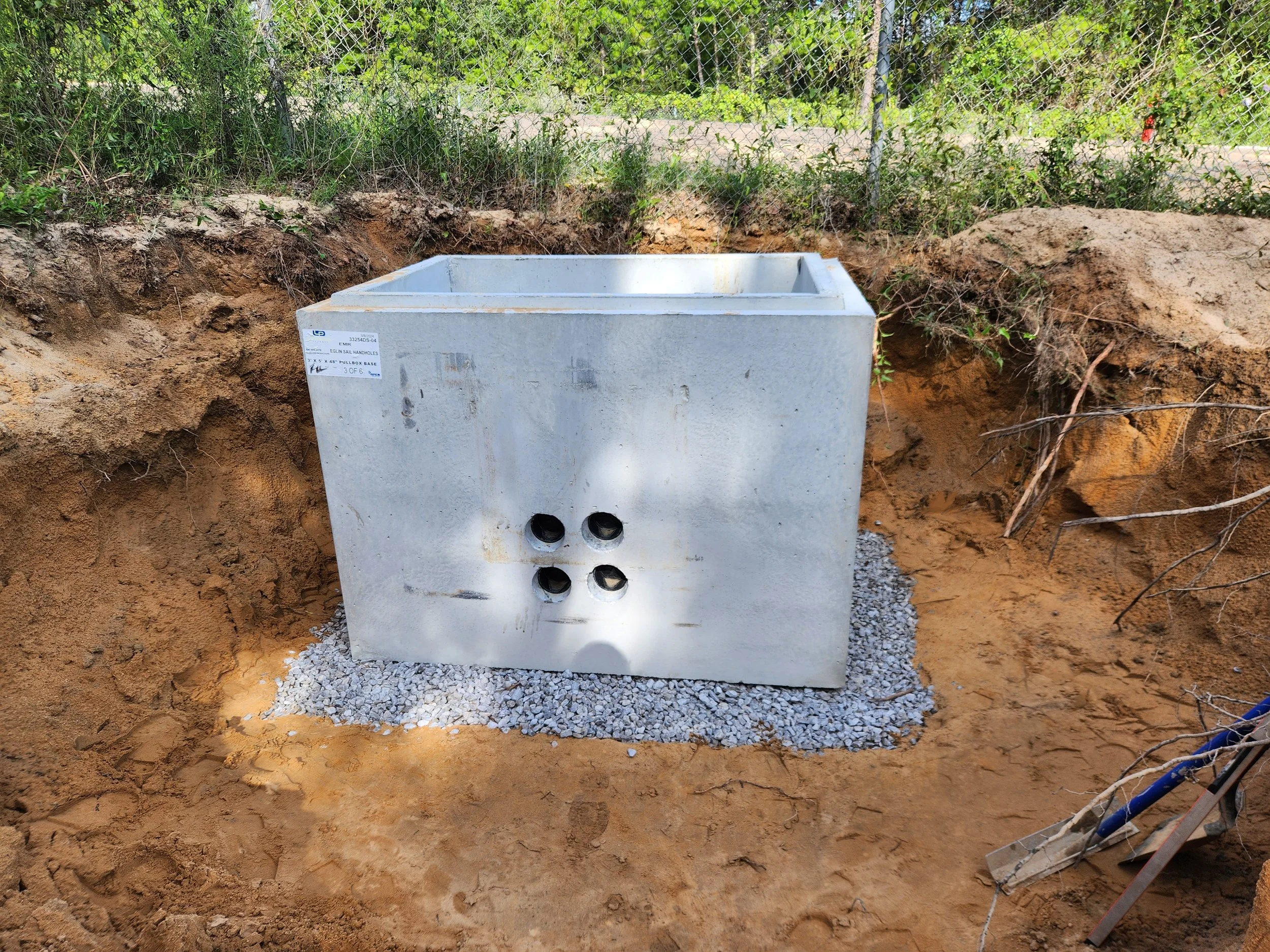 Underground utility valve box at a construction site, installed in a gravel bed with surrounding dirt and an excavated area, with green foliage and a chain-link fence in the background.