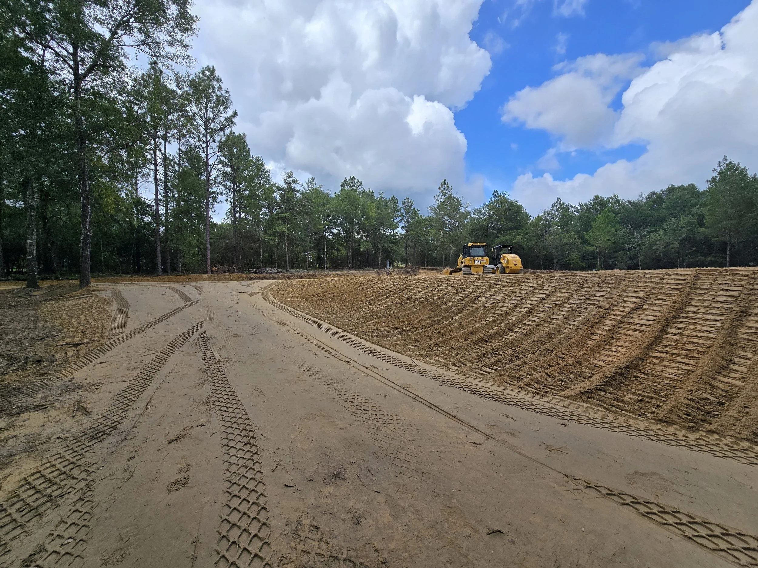 Construction site with a dirt road and two yellow bulldozers, surrounded by trees and under a partly cloudy sky.
