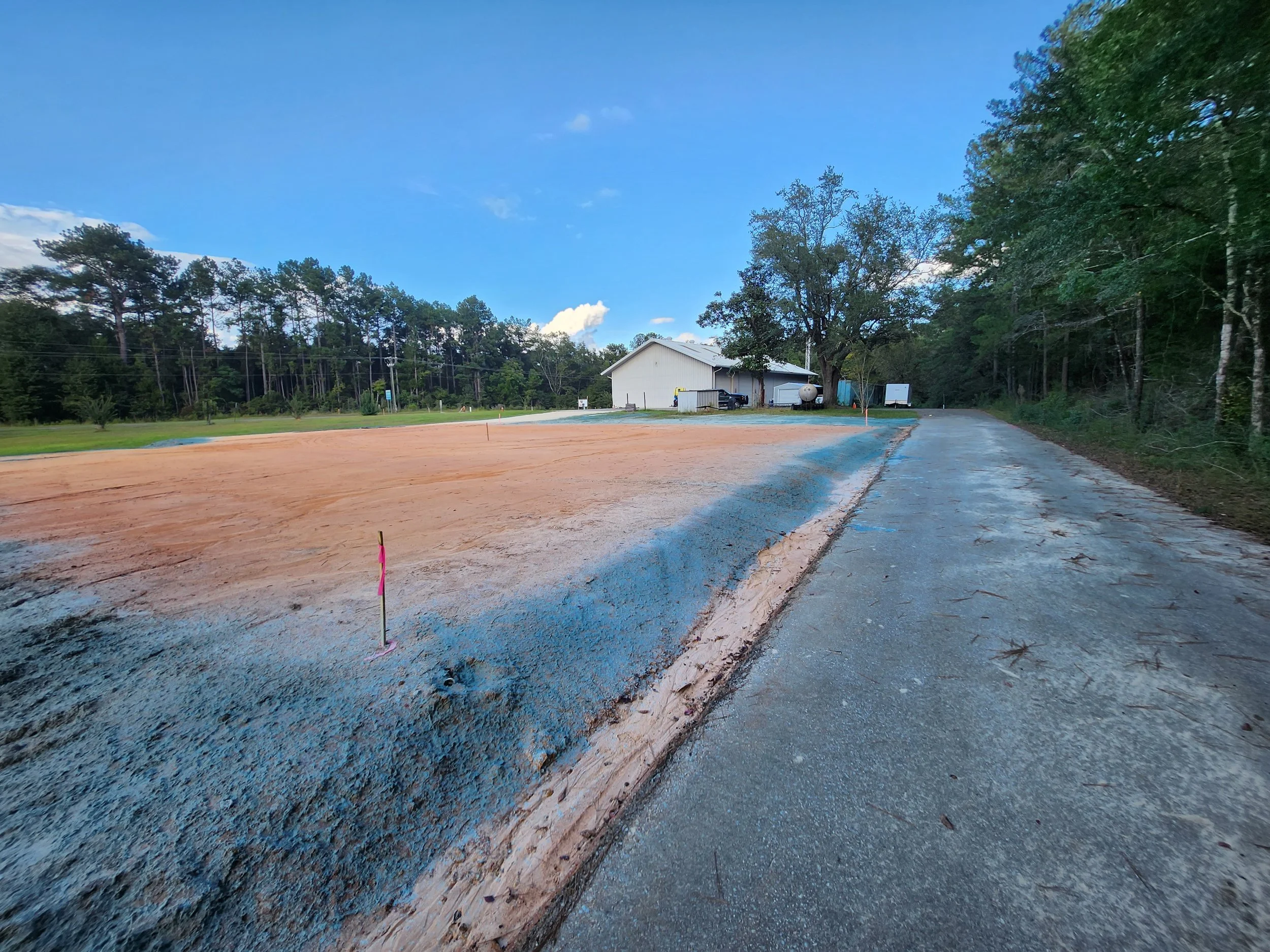 Construction site with gravel and dirt, with a building in the background, trees on both sides, and a partly cloudy sky.