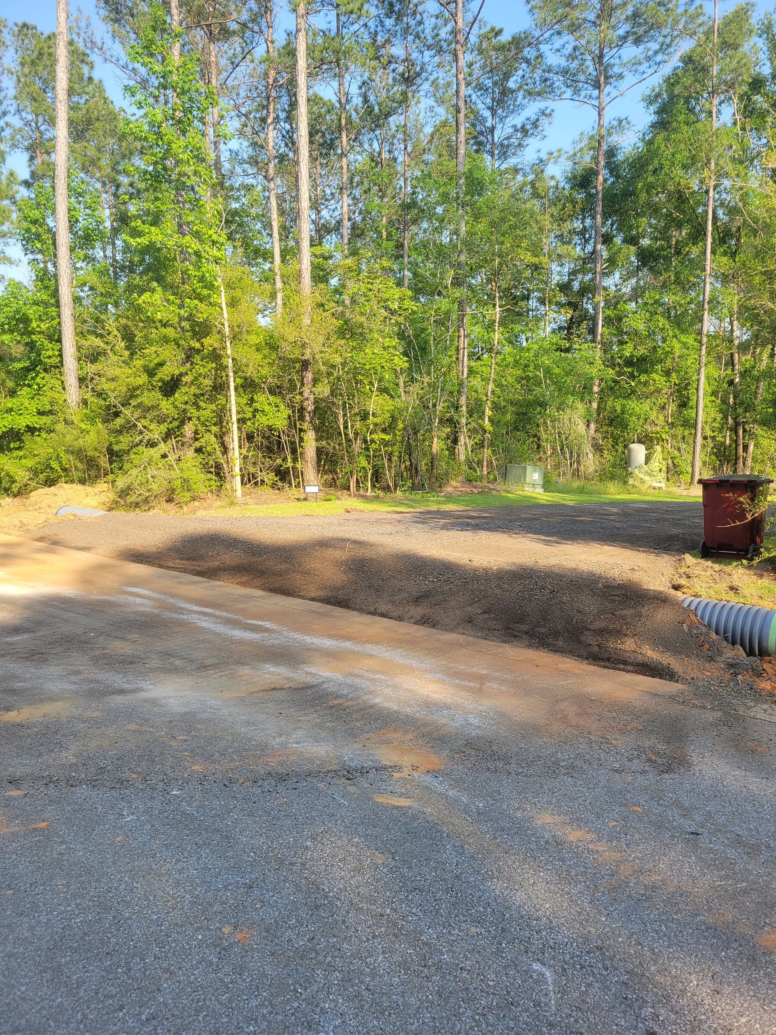 A construction site with a gravel and asphalt surface, surrounded by trees and utility boxes, with a red trash bin on the right.