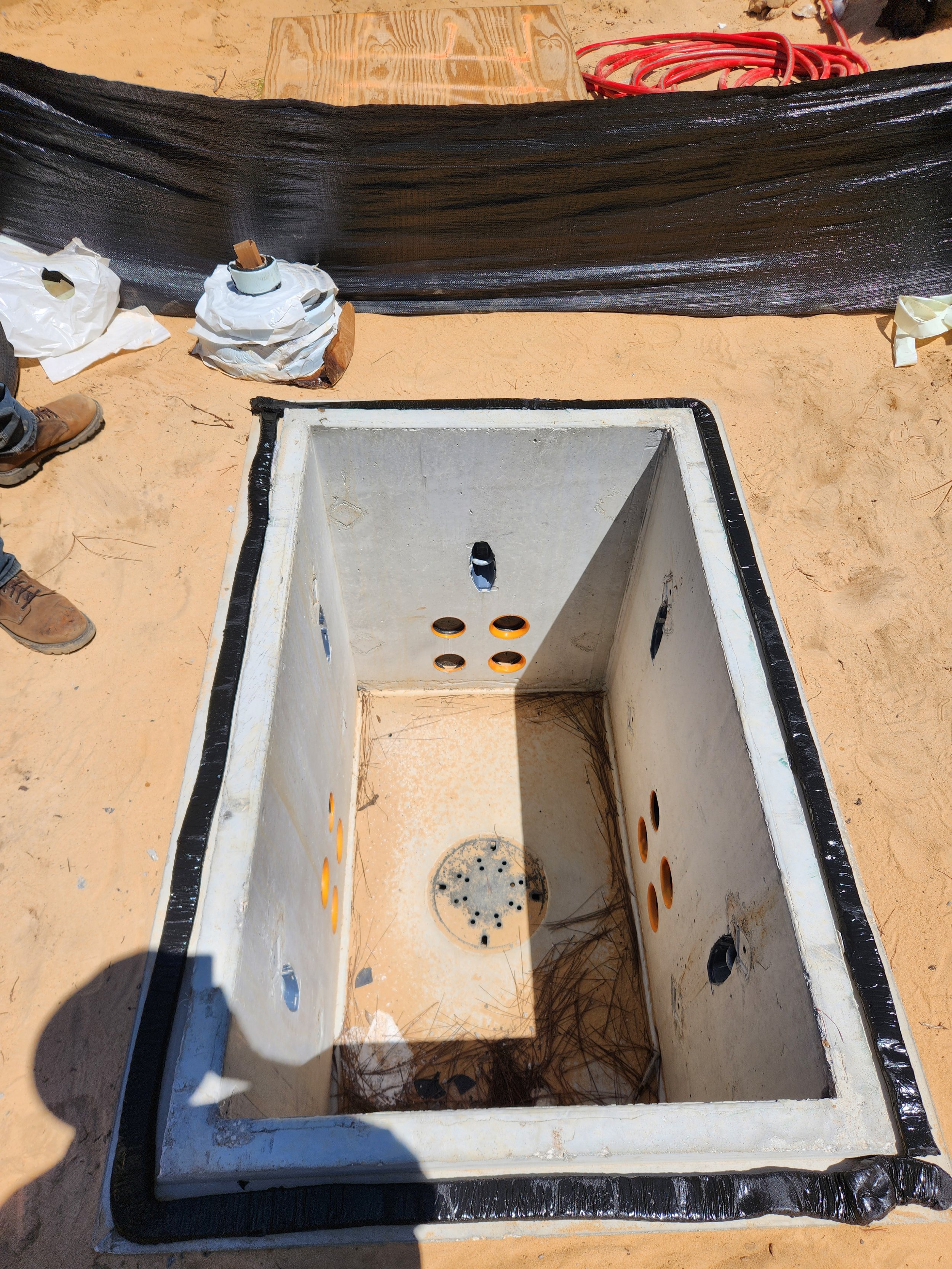 A rectangular concrete box with multiple circular holes on its sides and bottom, installed in sandy ground at a construction site. Black waterproofing material applied around the edges.