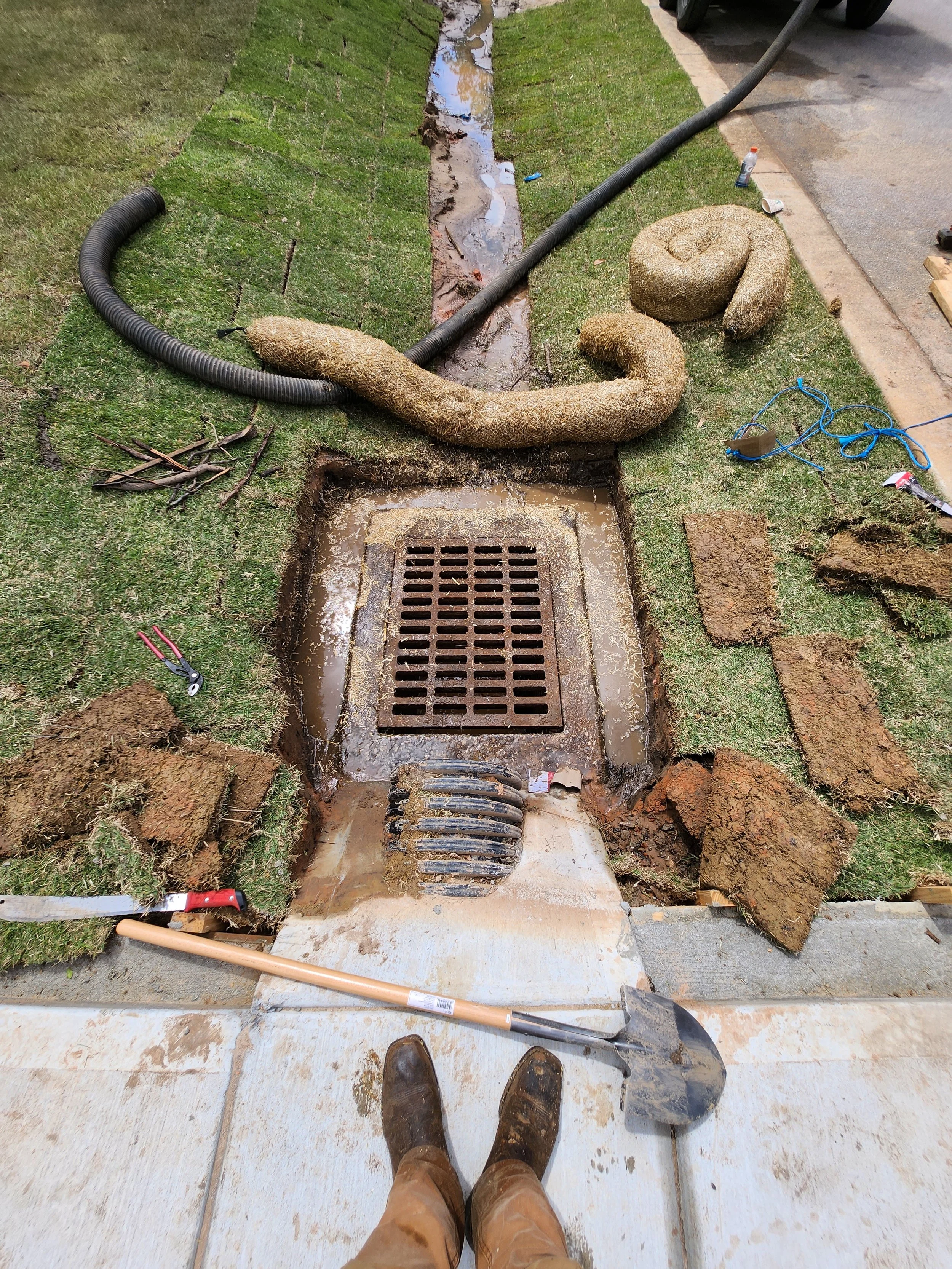 Construction worker standing in front of an open drain with tools and new drain tiles scattered around.