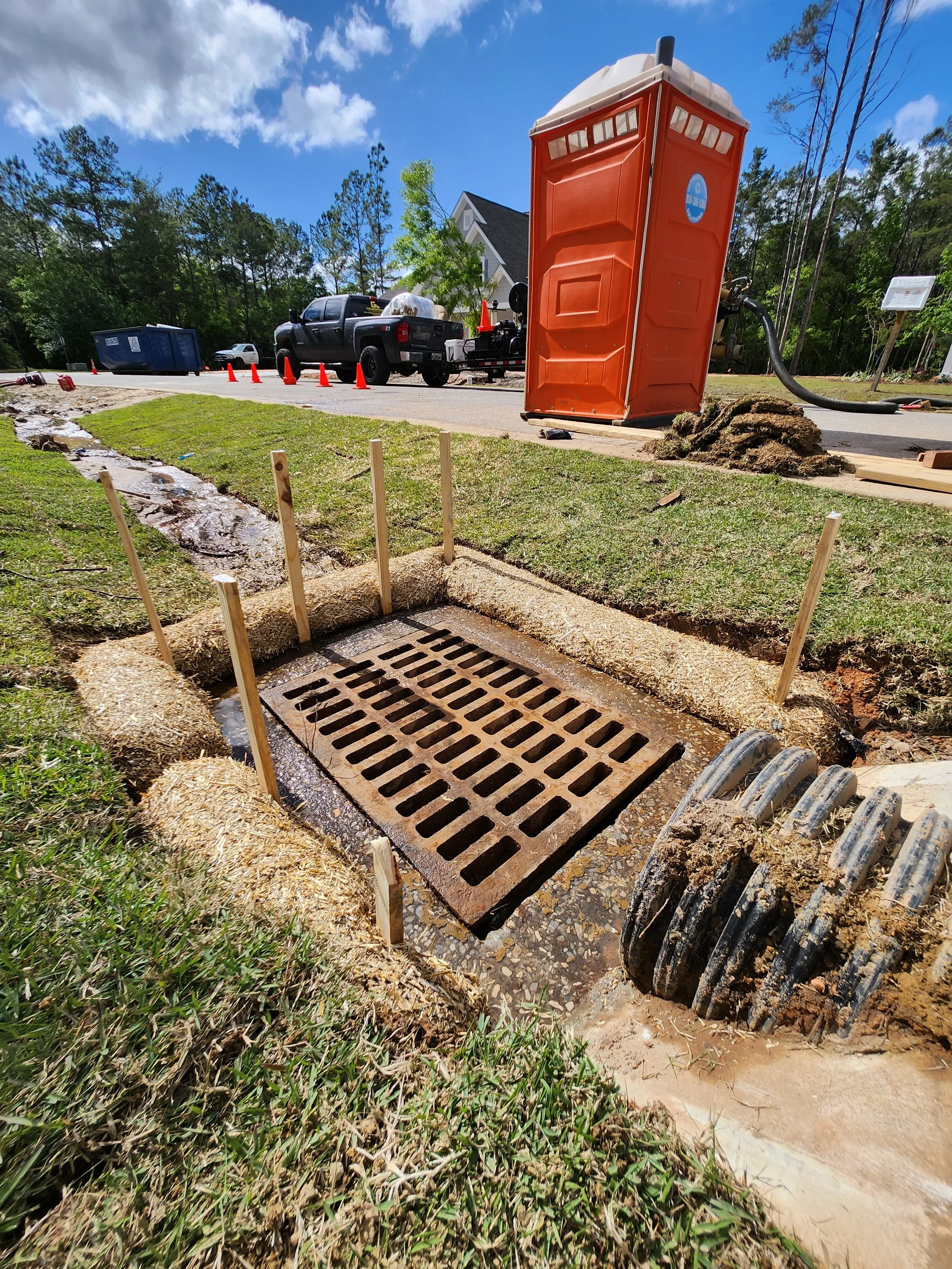 Construction workers installing a storm drain in a grassy area beside a street. A portable toilet and construction equipment are visible in the background.