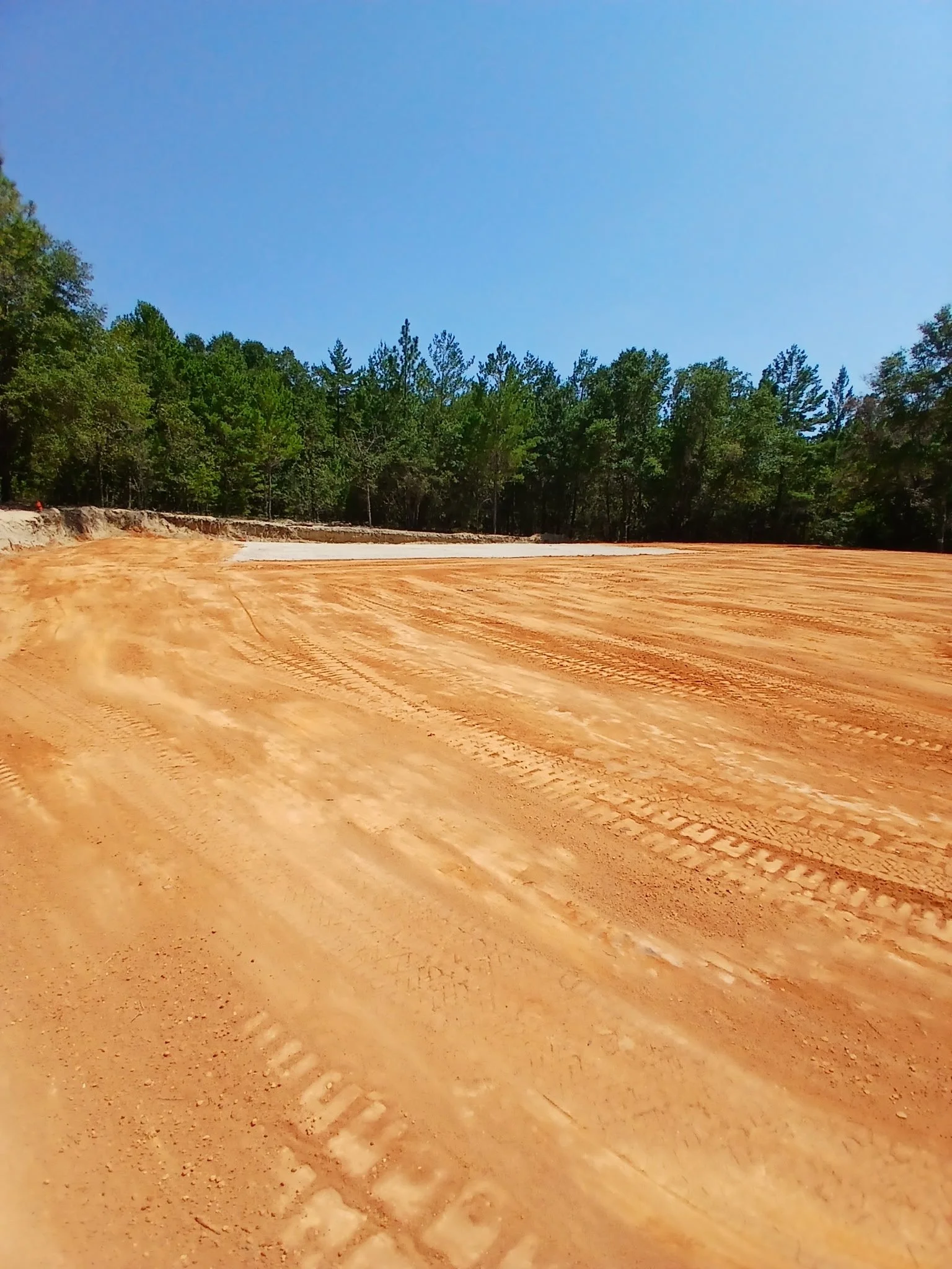 Dirt construction site with tire tracks and trees in the background under a clear blue sky.