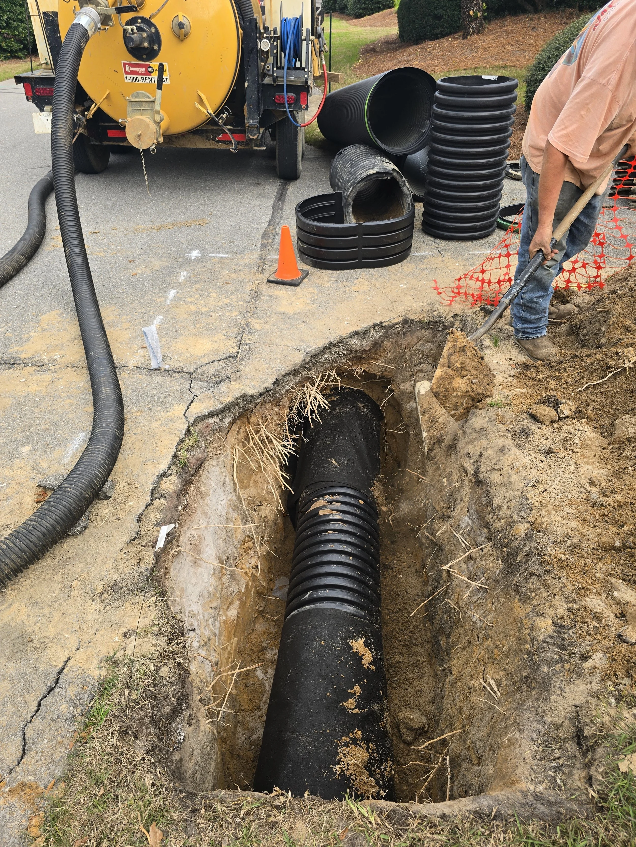 Worker installing or repairing underground drainage pipe using a shovel next to a trench with large black pipe sections and a vacuum truck nearby.