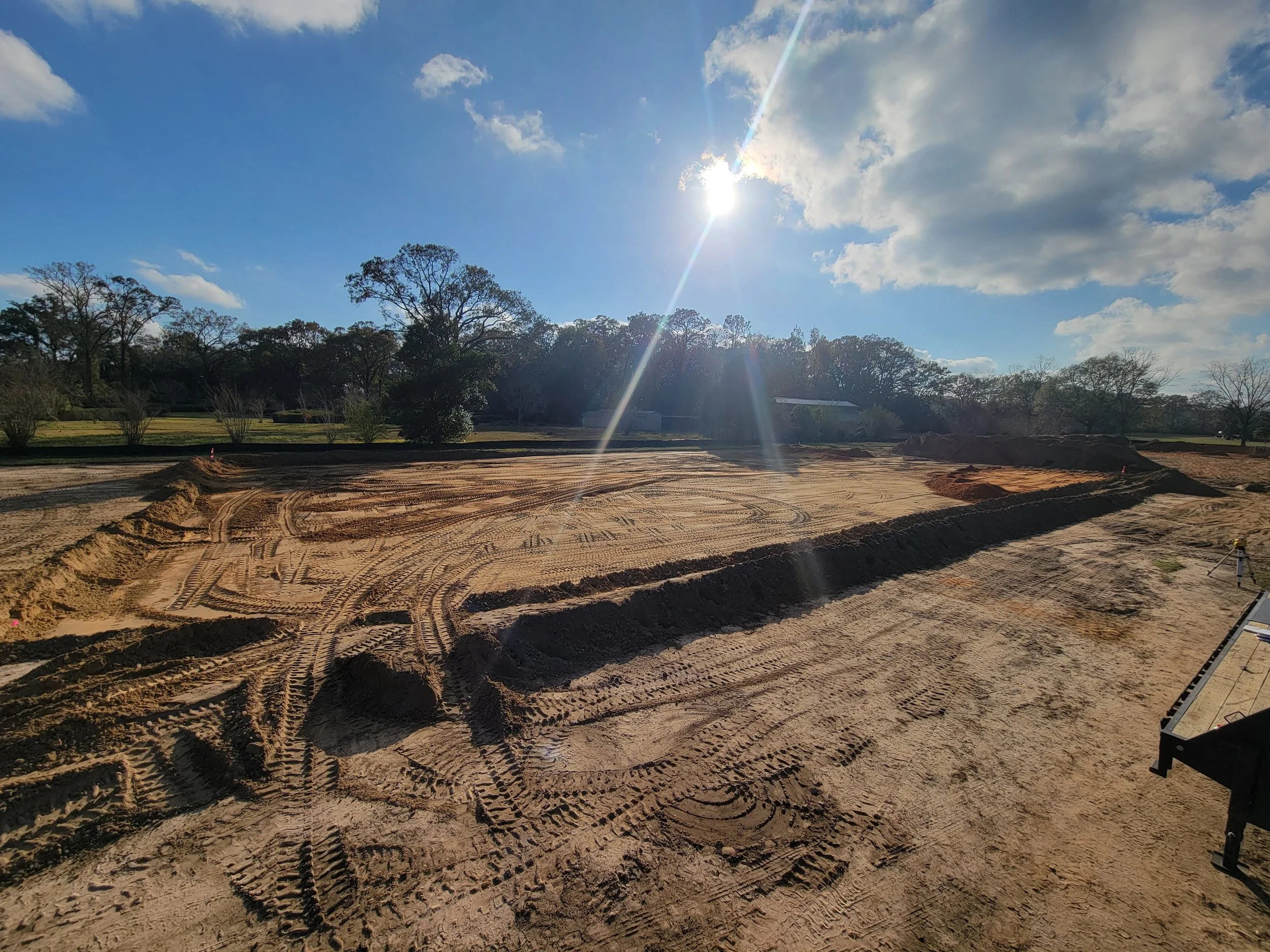 Construction site with freshly graded dirt and tire tracks, cleared for building, with trees and a blue sky with clouds and the sun visible overhead.