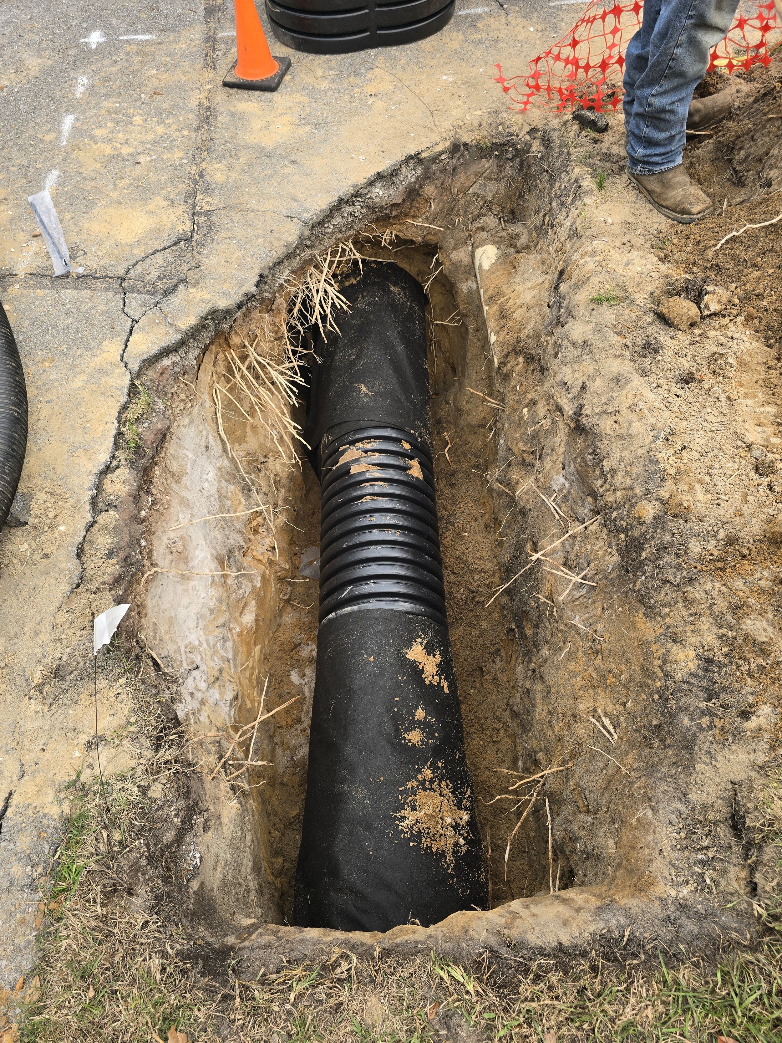 A construction site with a large black pipe in a trench, surrounded by dirt and soil. An orange safety cone and a person wearing jeans and work boots are visible nearby. There is an orange plastic safety fence nearby.
