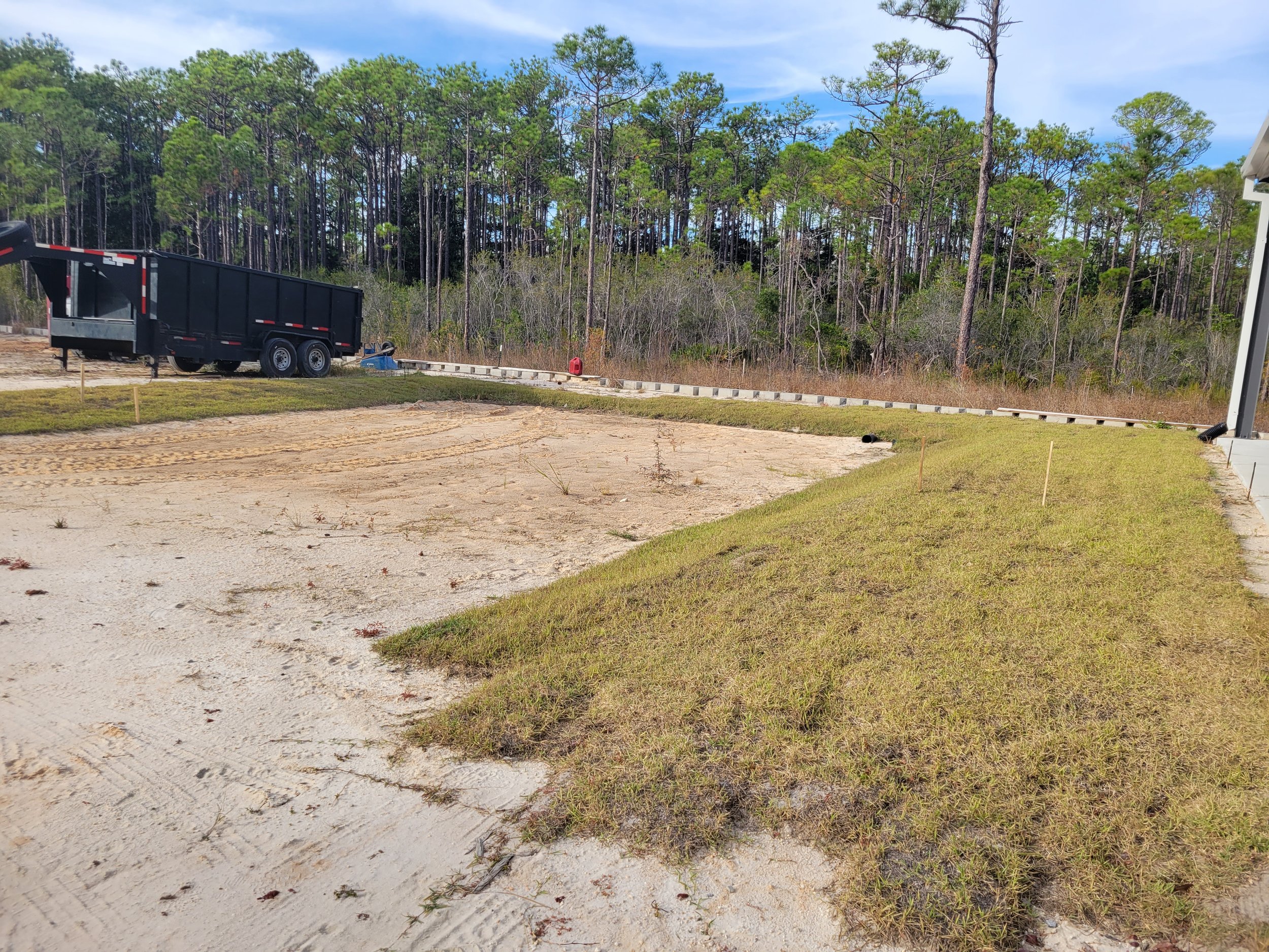 Construction site with a black dump trailer and a grassy area in the foreground, sandy ground, trees in the background, and a partly cloudy sky.