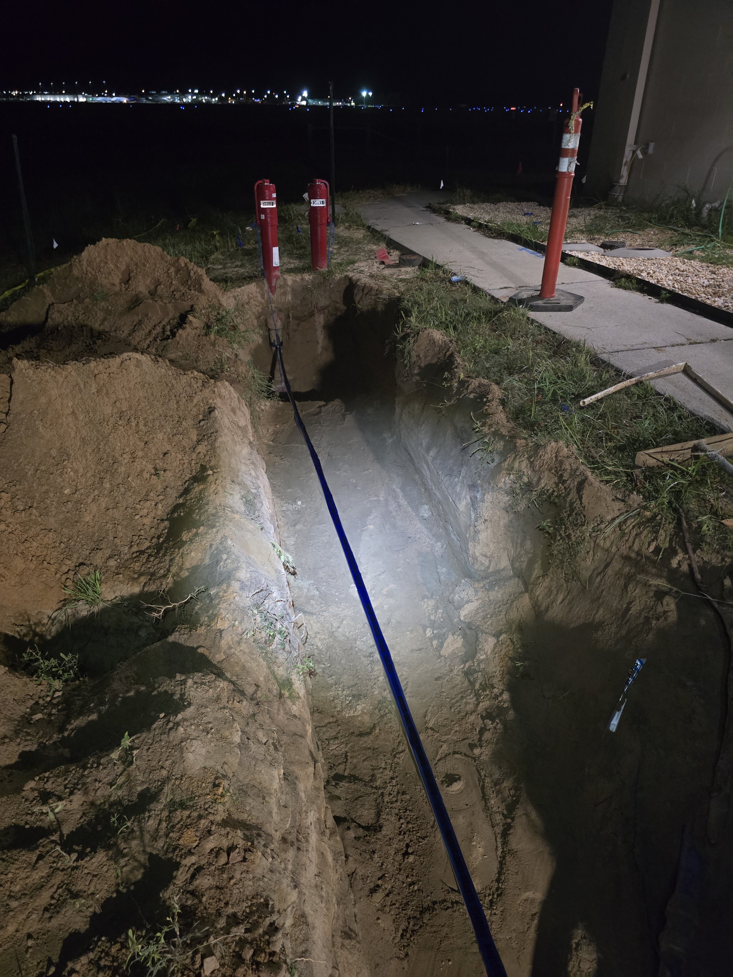 Nighttime construction site showing an excavated trench with a blue cable or pipe inside, surrounded by soil and construction equipment, with a sidewalk, orange cones, and fire extinguishers nearby