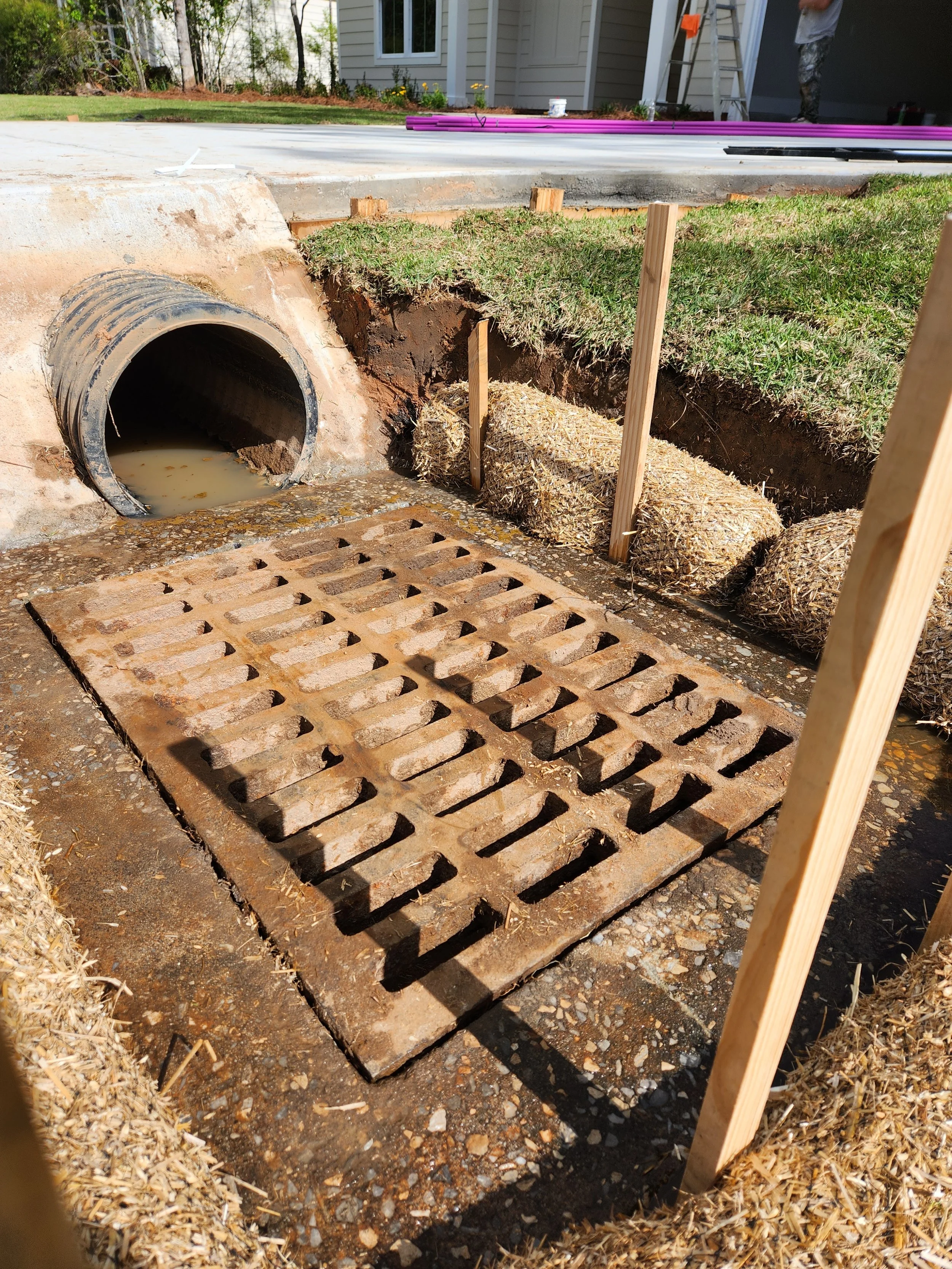 Construction site showing a drain grate installed over an opening with a pipe and hay bales supported by wooden stakes beside excavation.