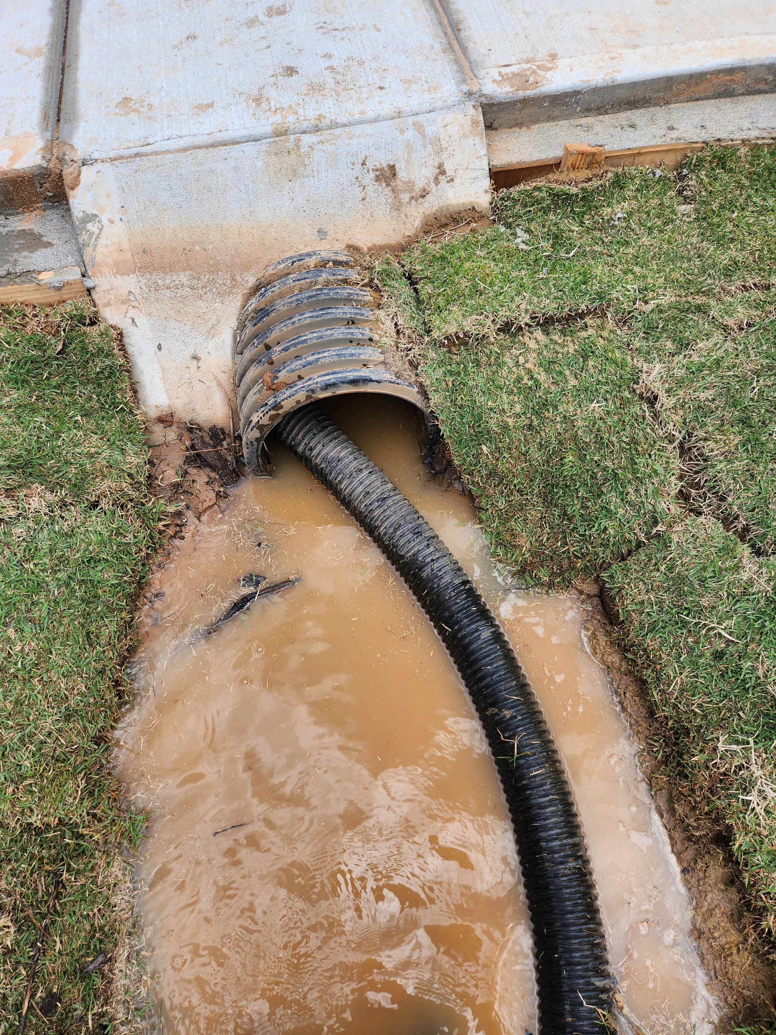 Drain pipe leading into a dirt and grass trench filled with muddy water.