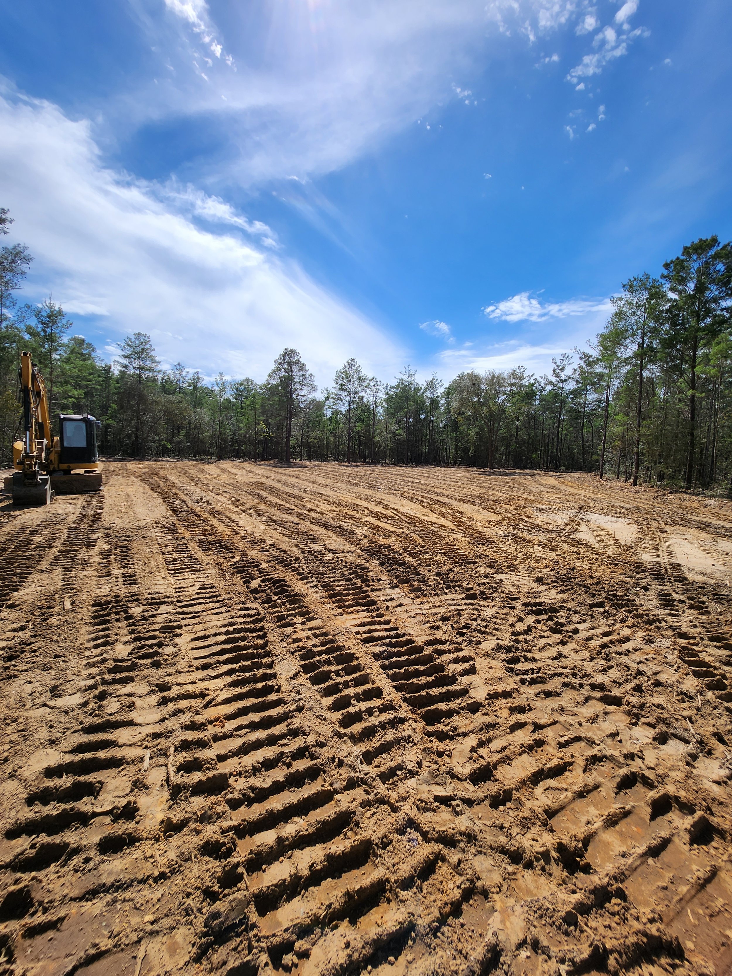 Empty construction site with freshly dug dirt, a yellow excavator in the background, and a partly cloudy sky.