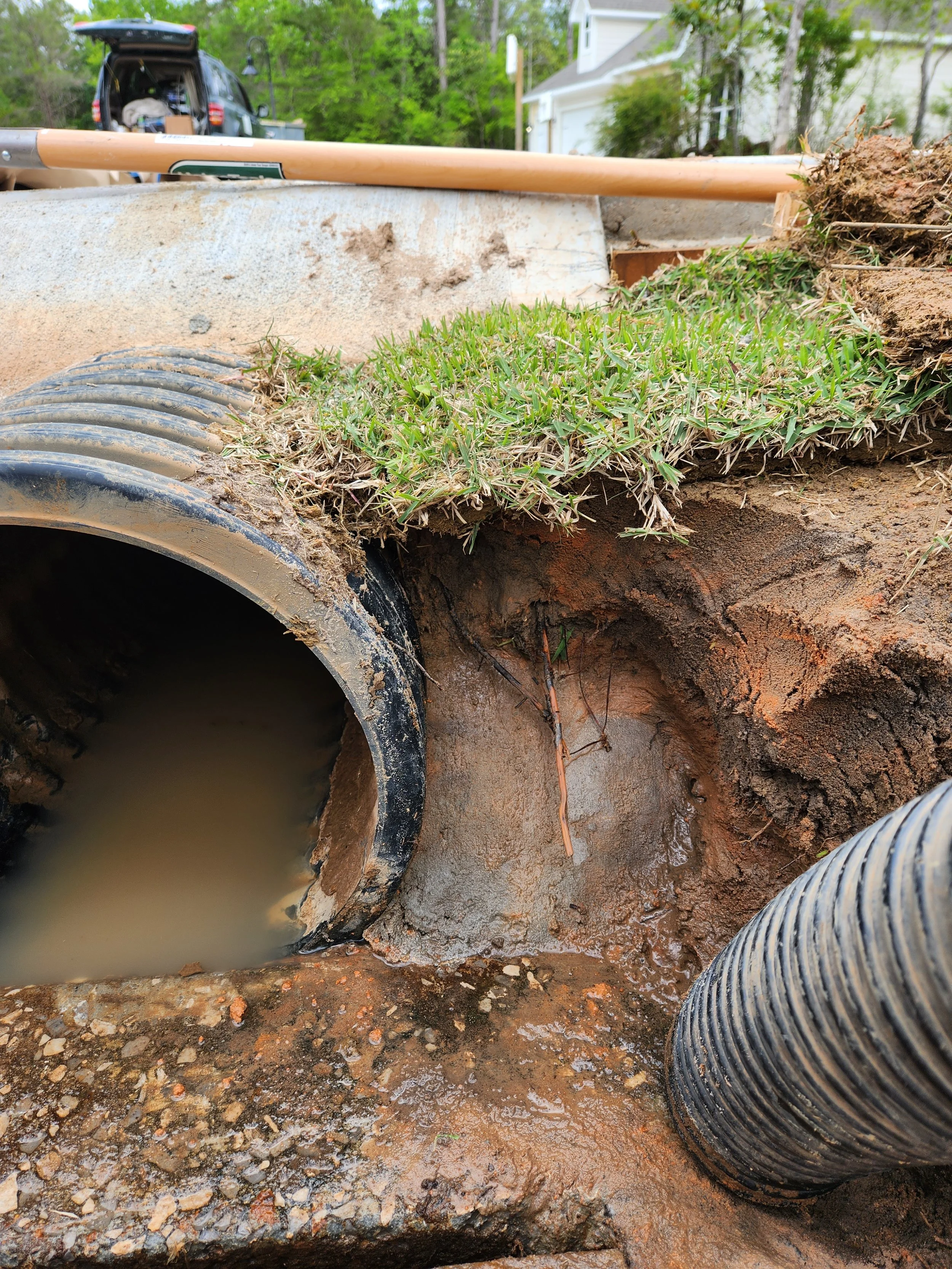 Underground pipe installation in progress with water around the pipe, a grassy soil surface above, and construction tools and vehicles in the background.