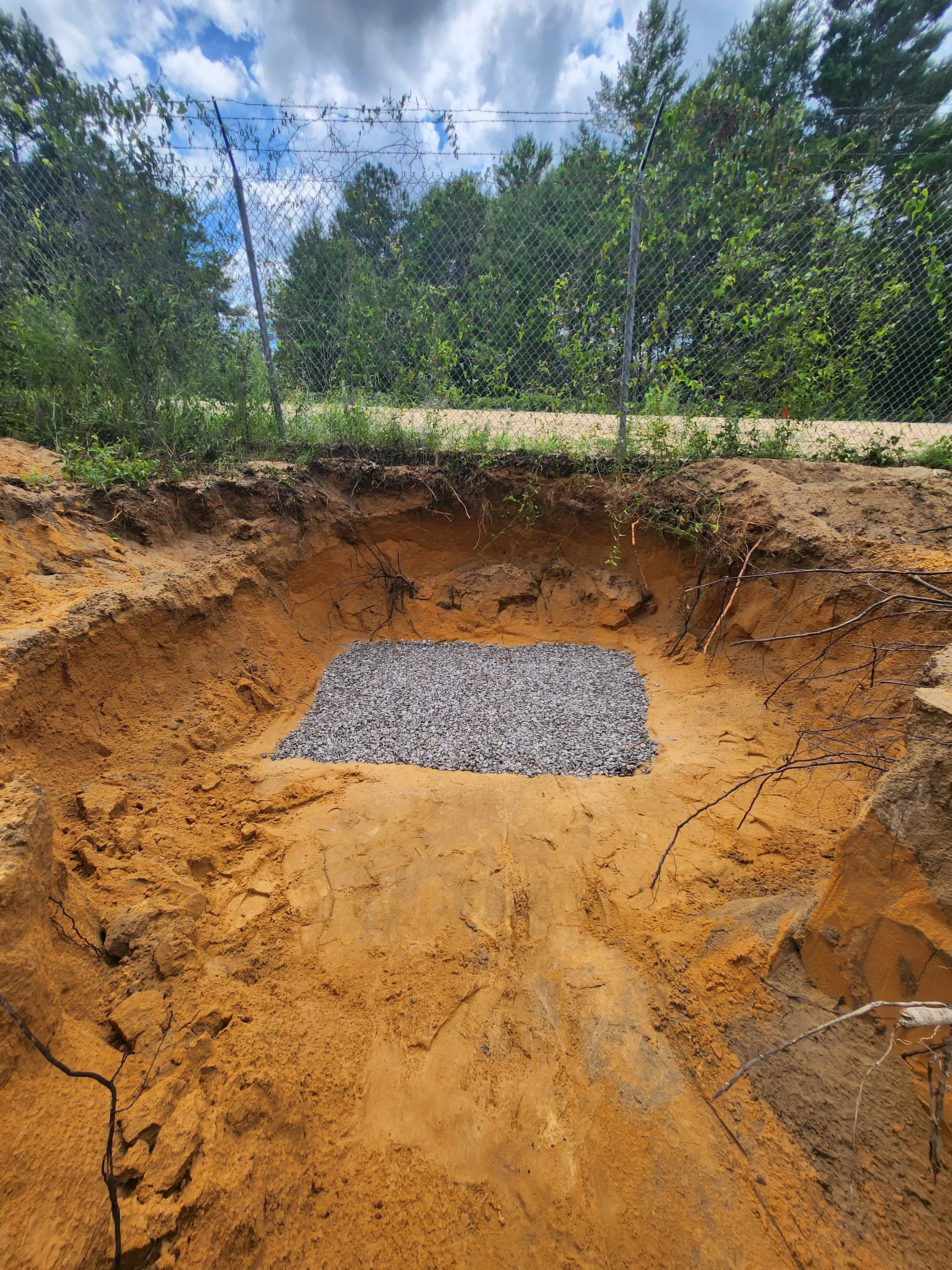 A freshly dug hole in the ground with a layer of gravel placed at the bottom, surrounded by earth and grass, with a chain-link fence and trees in the background.