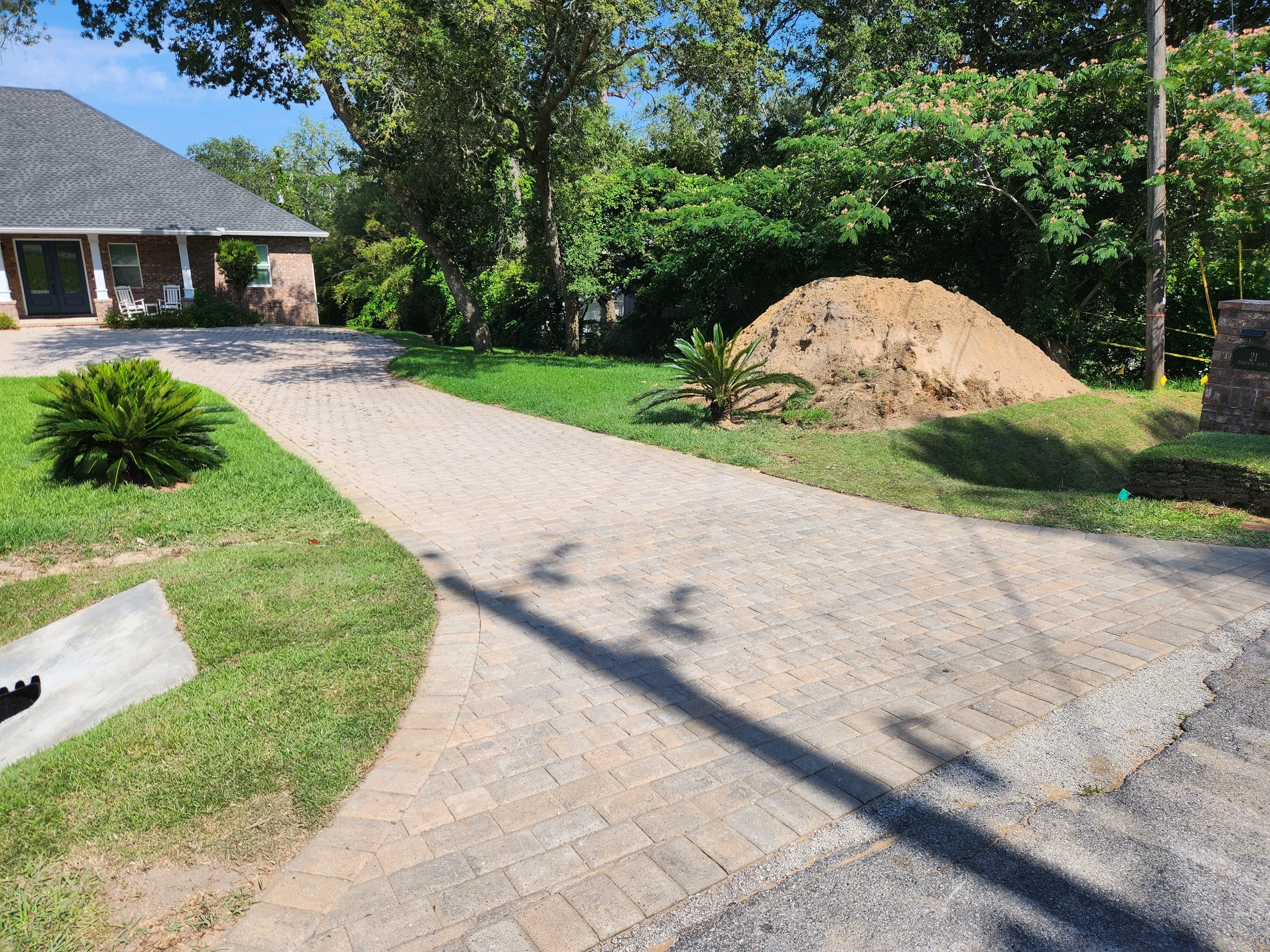 A newly paved brick driveway in front of a brick house with a front porch and white chairs. There are small plants and trees along the driveway, with a large tree and a pile of dirt on the lawn, and a brick mailbox near the edge of the driveway.