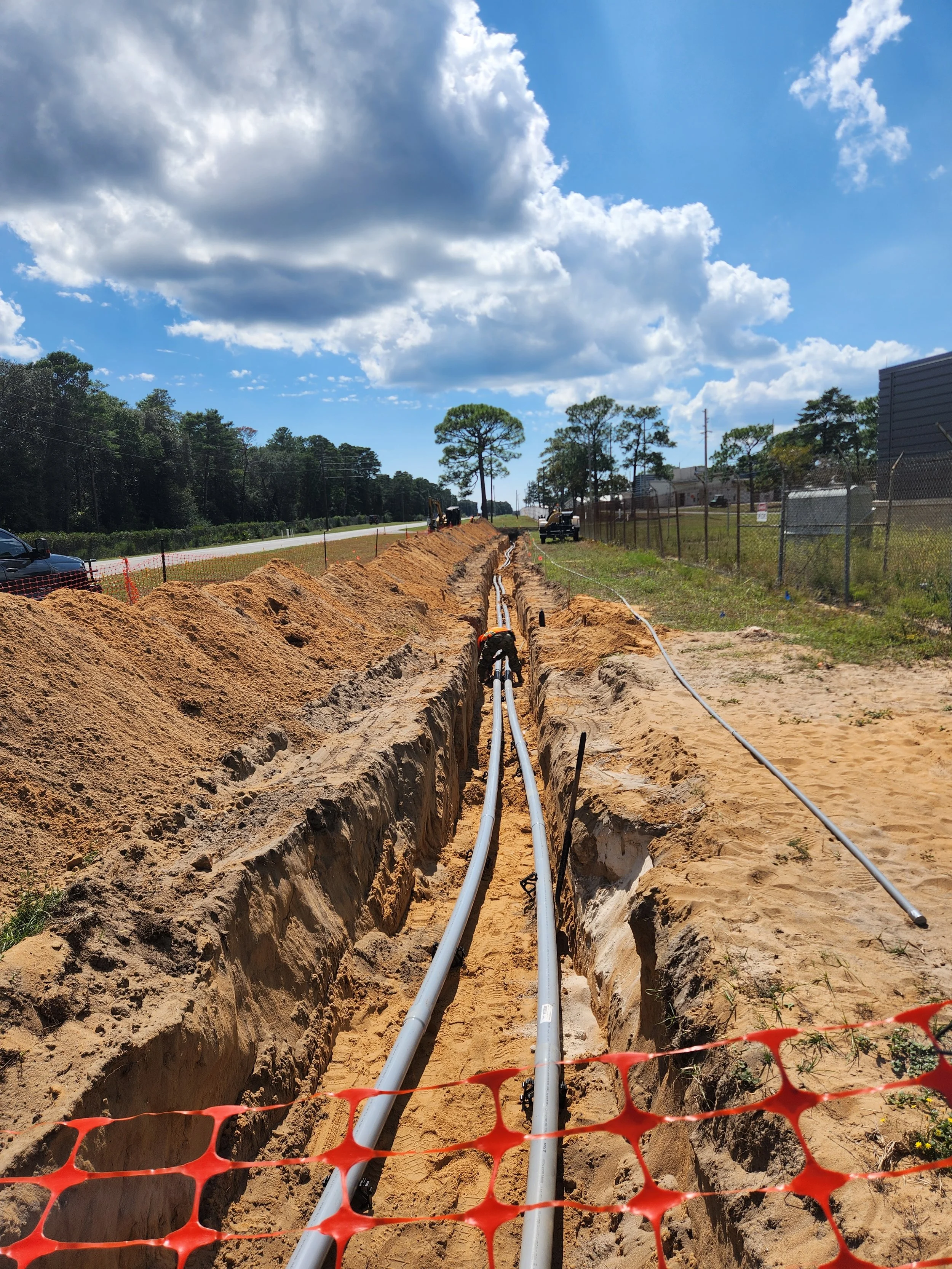 Construction site with trenches and pipeline installation on sandy soil, with orange safety fencing and construction equipment in the background, under partly cloudy sky.
