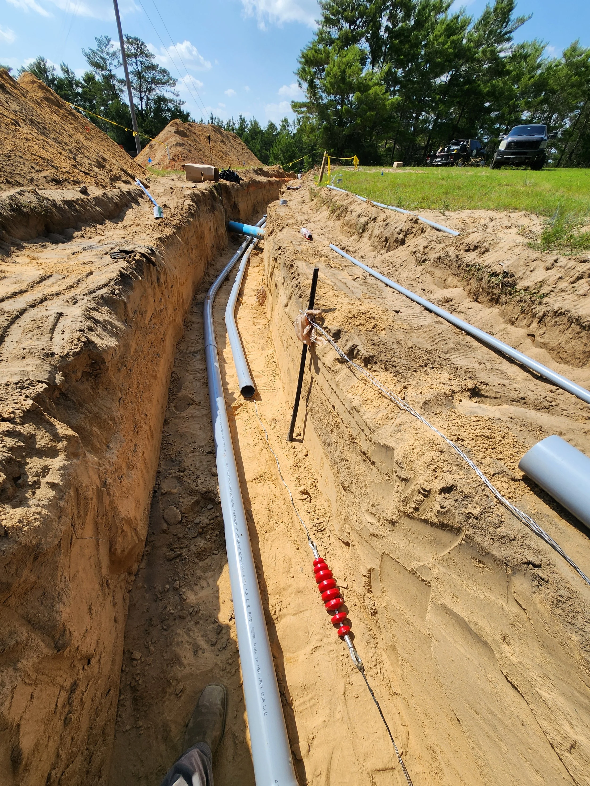 Construction site with trenches dug and pipes installed underground, with a clear sky and trees in the background.