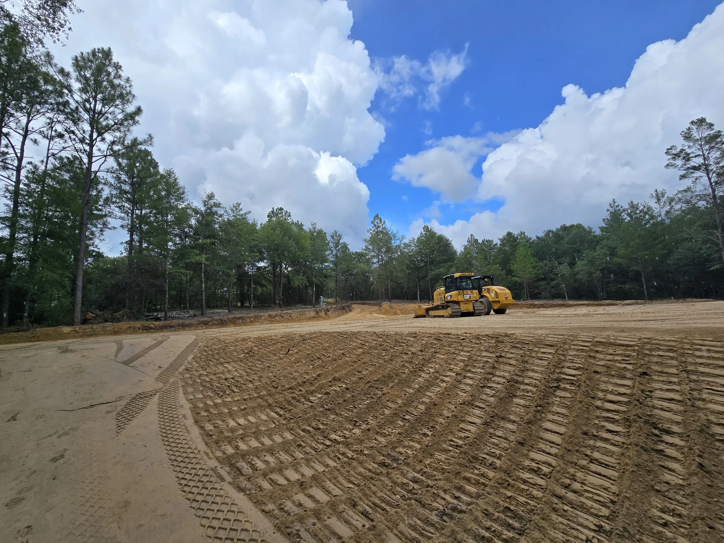 A construction site with a yellow bulldozer working on the dirt ground under a partly cloudy sky, surrounded by trees.