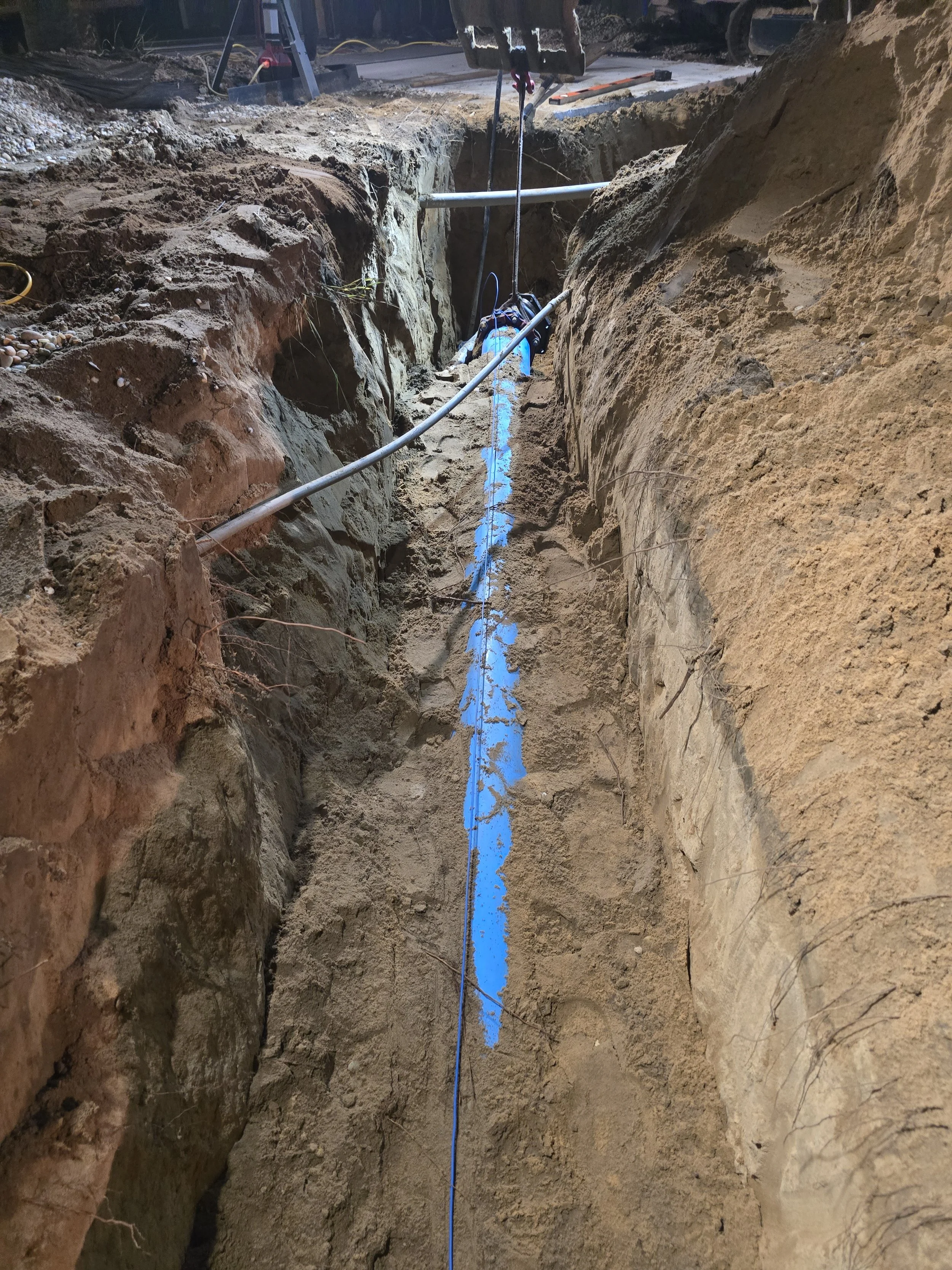 A construction site with a deep trench in the ground, showing soil layers on both sides. There is a blue line painted along the bottom of the trench, and various construction tools are visible at the top.