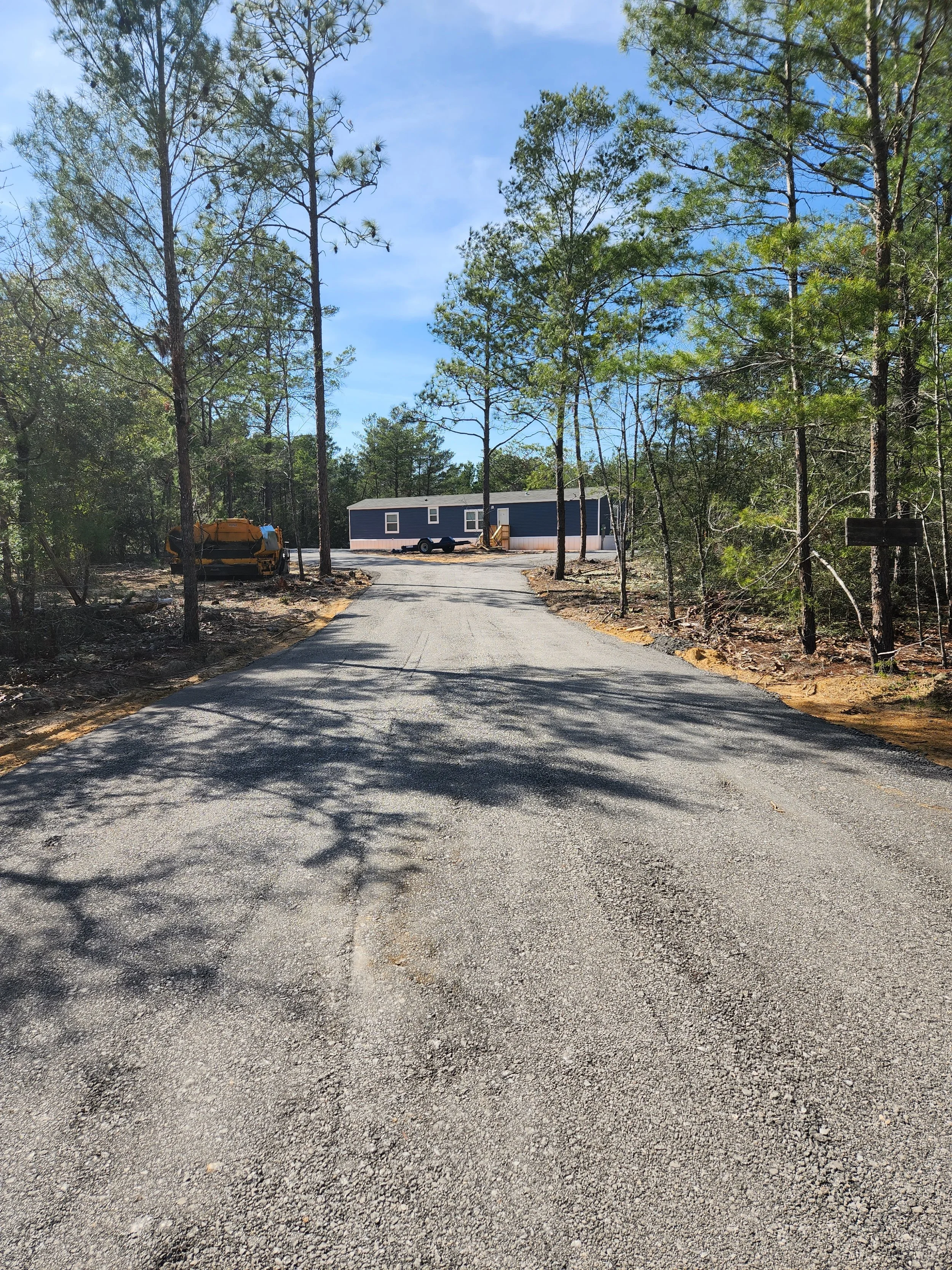 A newly paved gravel road leading to a small blue house with white trim, surrounded by trees and a trailer in a wooded area.