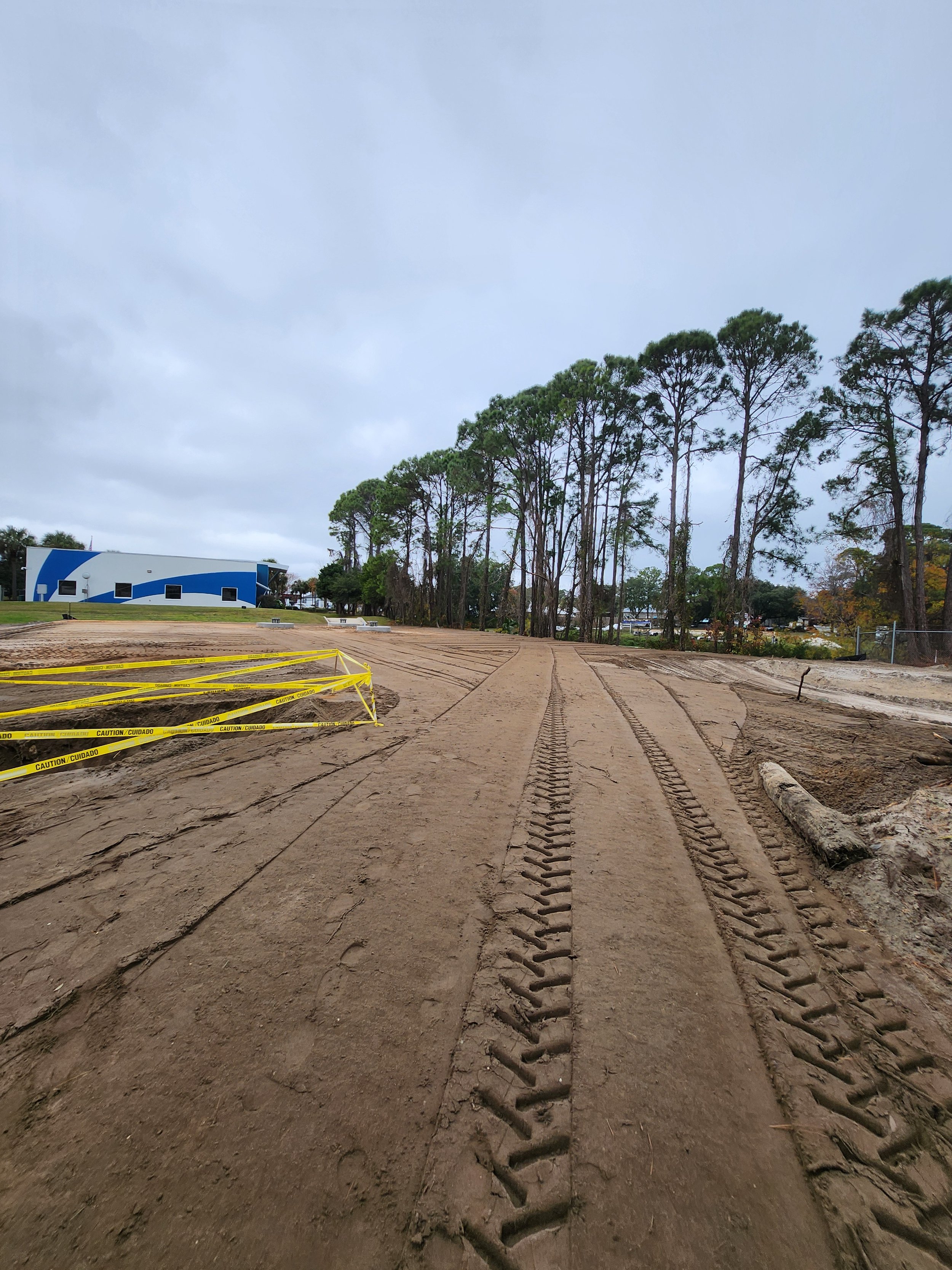 Construction site with freshly graded dirt, tire tracks, and yellow caution tape, surrounded by tall trees and a blue and white building in the distance under cloudy skies.