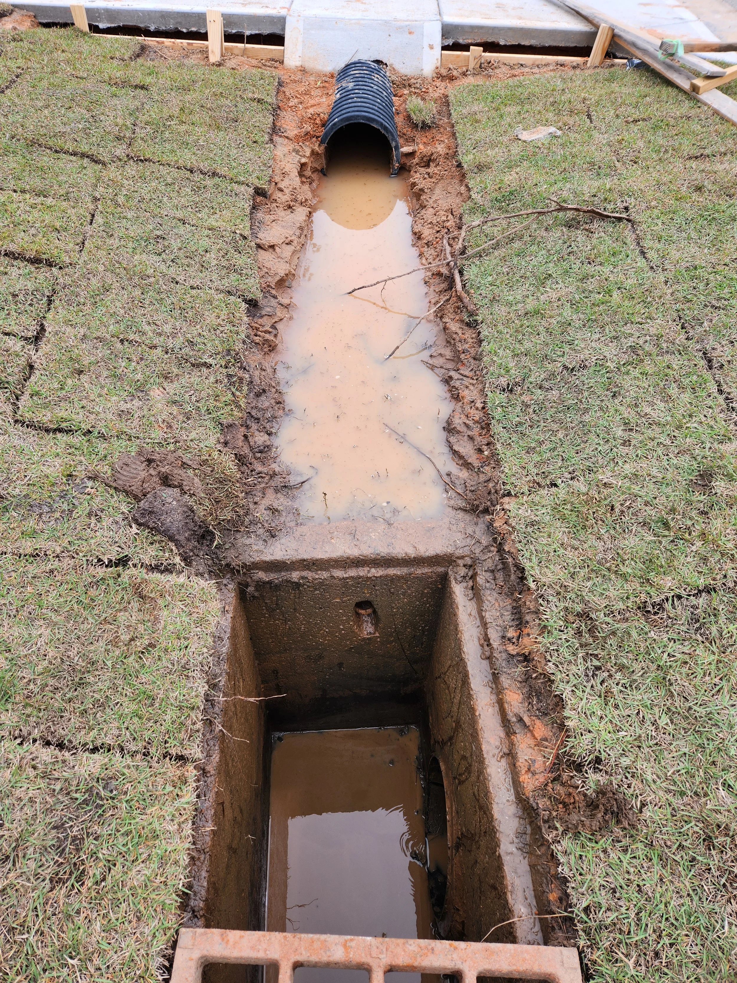 A construction site with a trench for underground piping, including a black corrugated pipe and two water-filled sections, surrounded by grass and some construction materials.