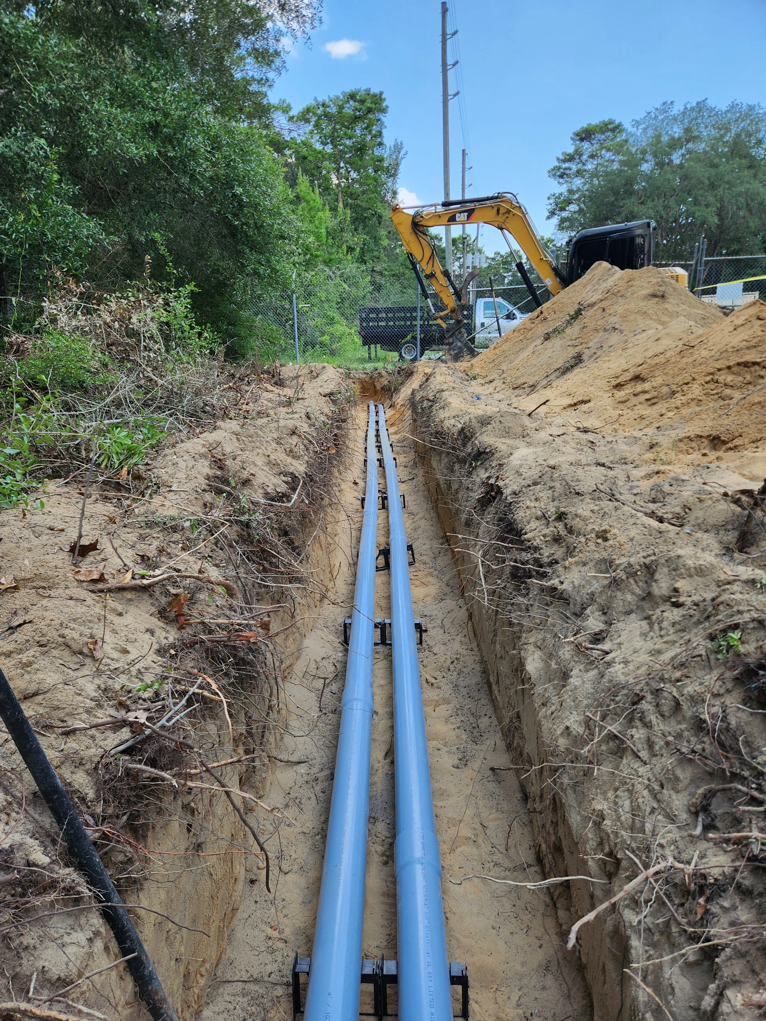 Two blue pipes installed in a trench with a yellow excavator nearby on a construction site surrounded by trees and utility poles.