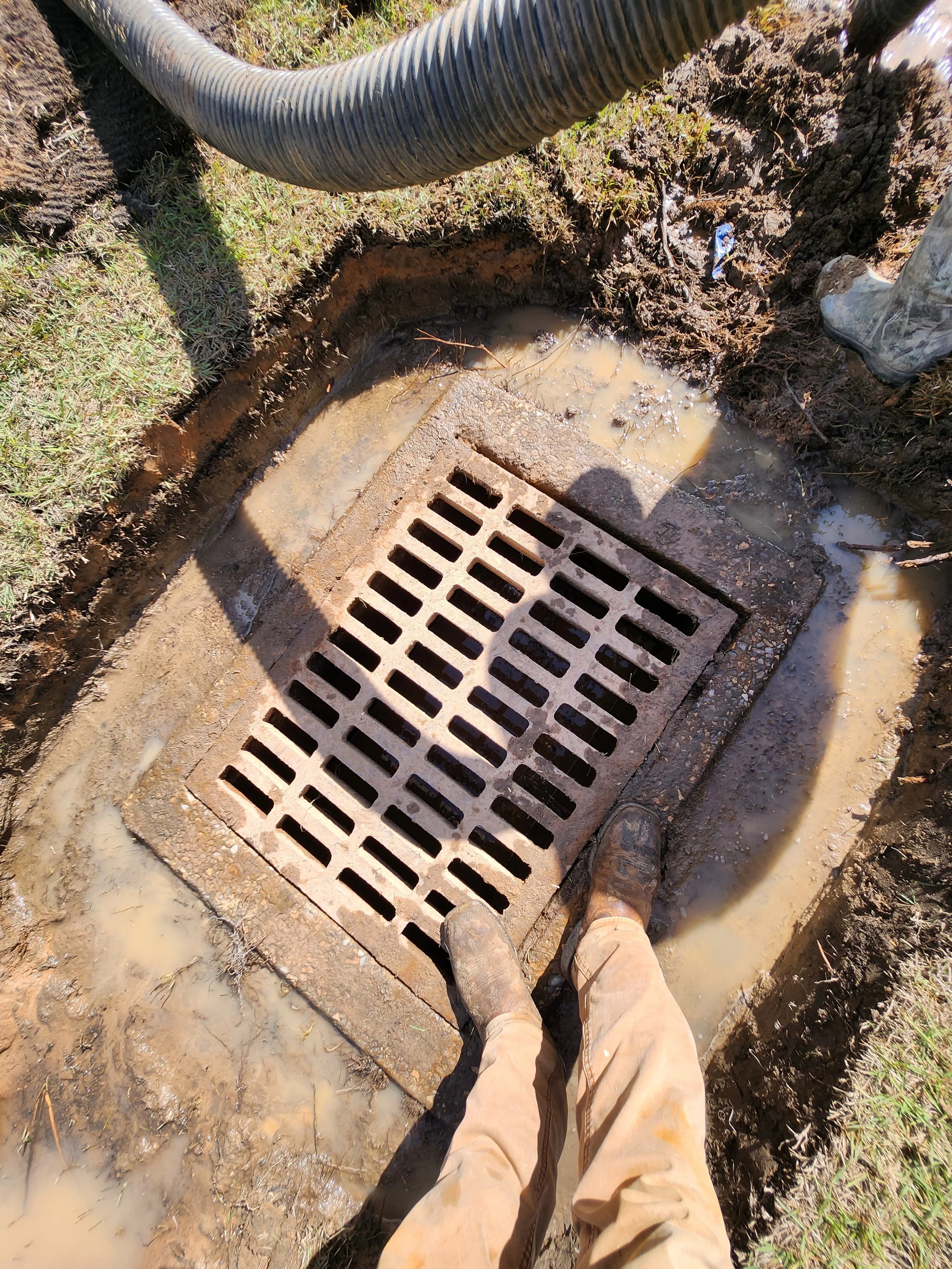 Person standing over an open storm drain in muddy water, wearing work boots and tan pants, with a black flexible pipe nearby.