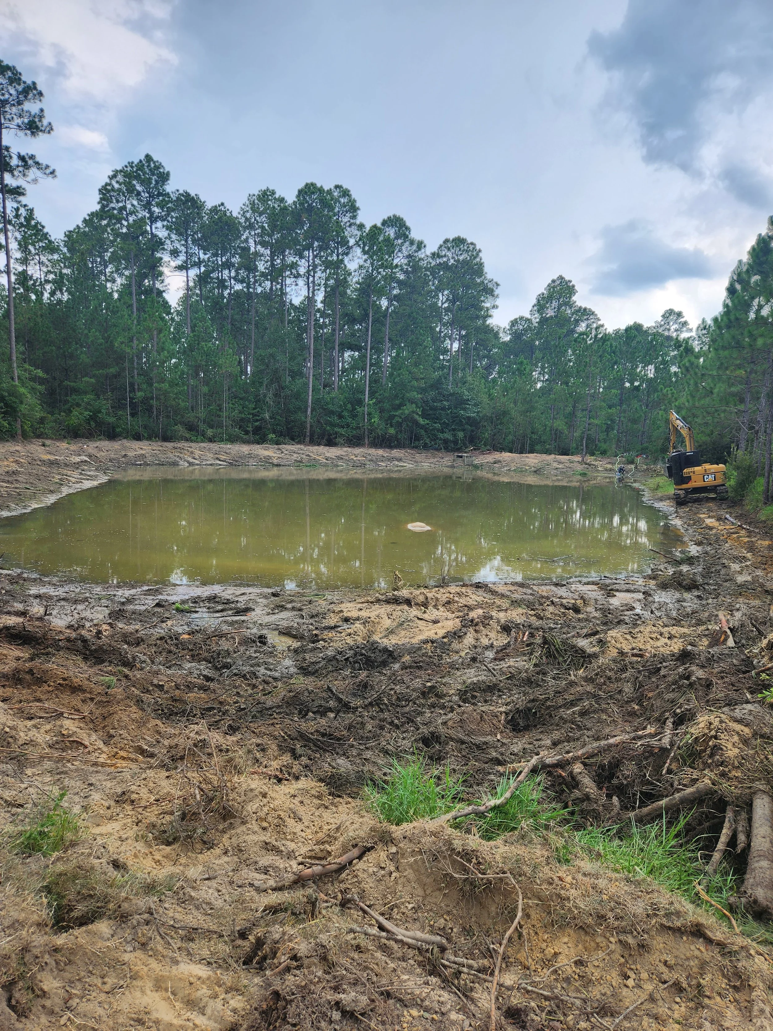 A small pond with muddy banks, surrounded by trees, with construction equipment on the right side.