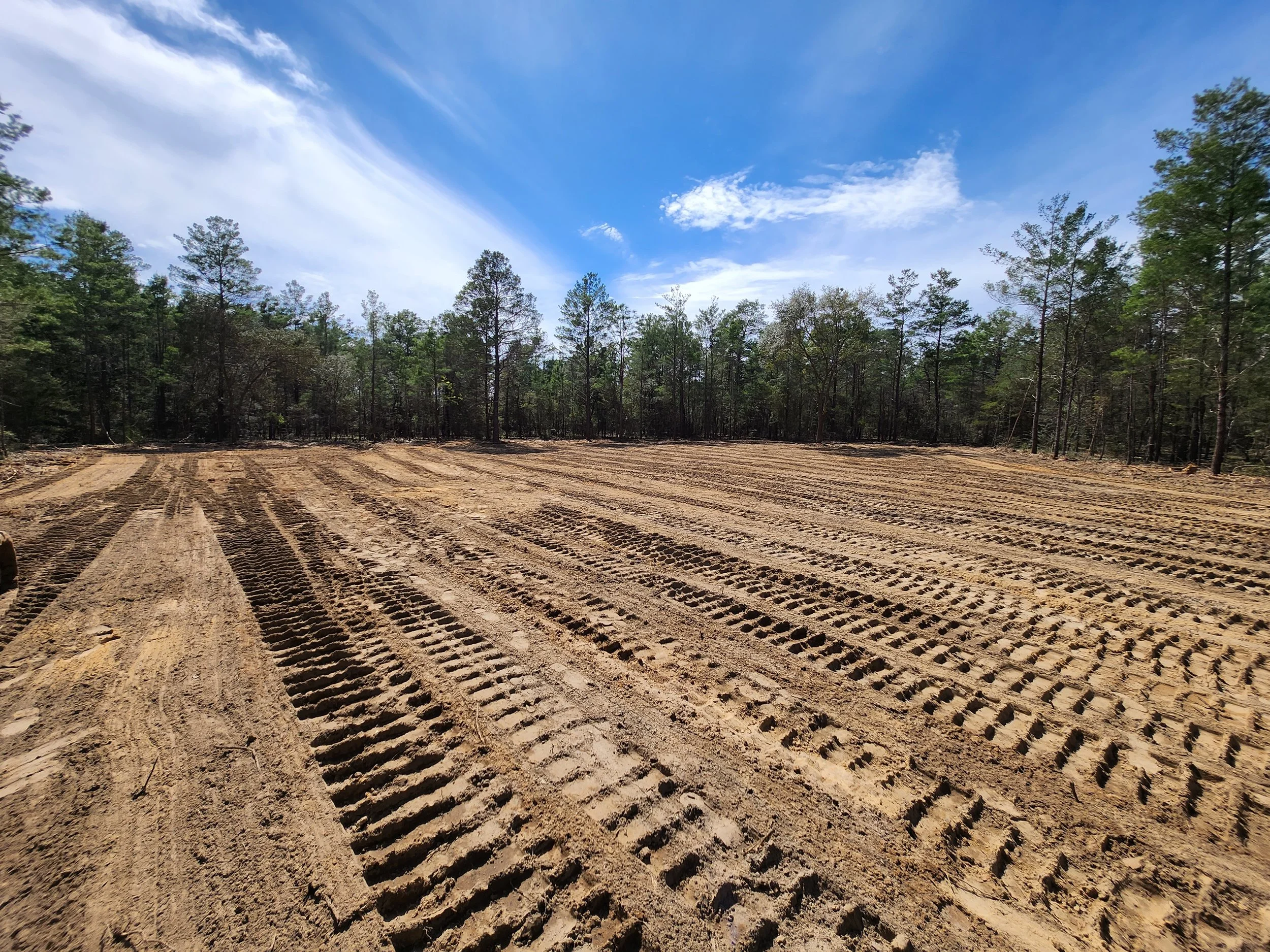 Freshly tilled soil in a cleared field with tire tracks, surrounded by trees under a partly cloudy blue sky.