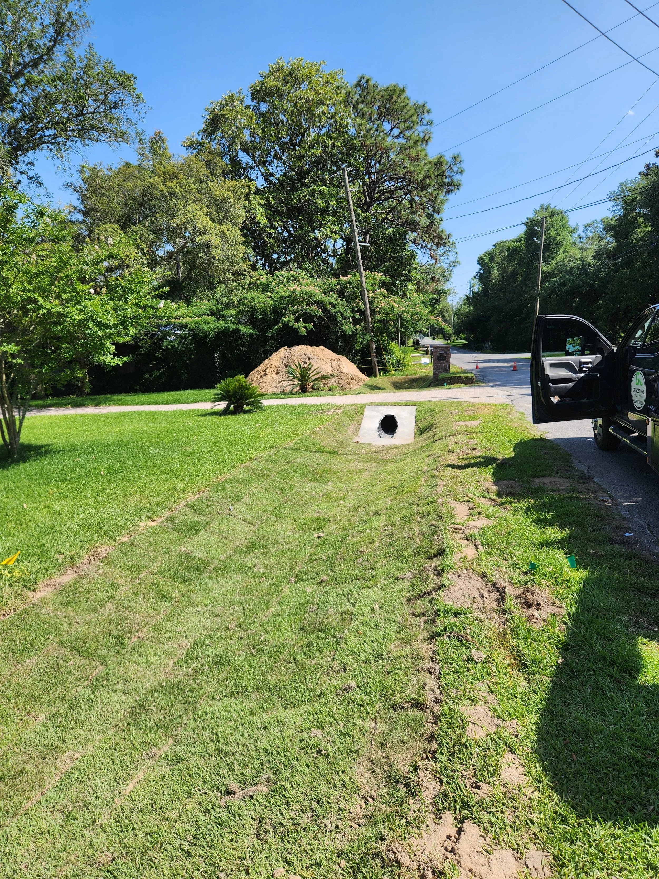 A suburban street with a construction pipe in the ground, a truck parked on the side, and a large dirt pile amidst green trees and grass under a clear blue sky.