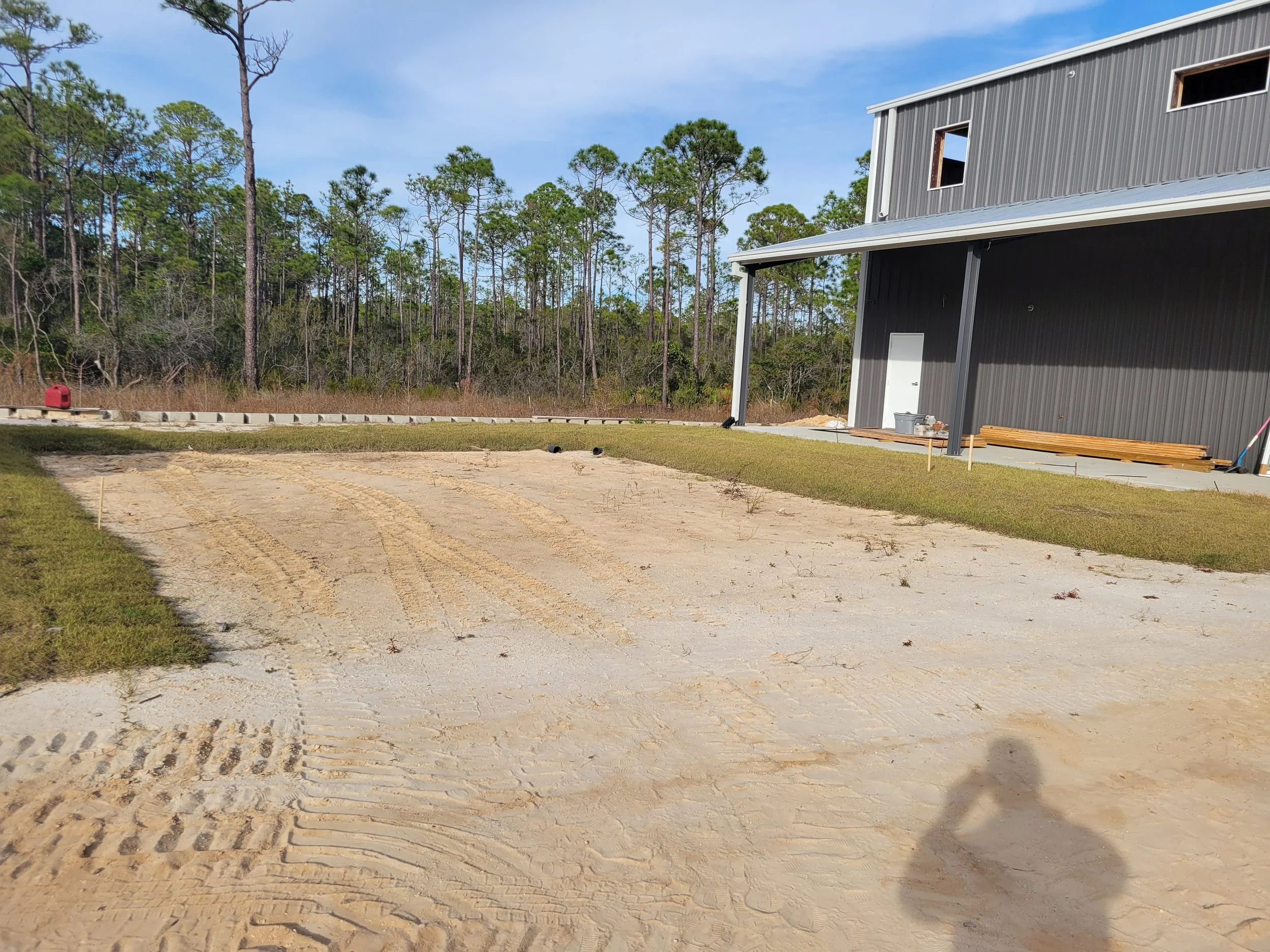 Construction site with a partially built house, dirt driveway with tire tracks, grass patches, and surrounding wooded area.