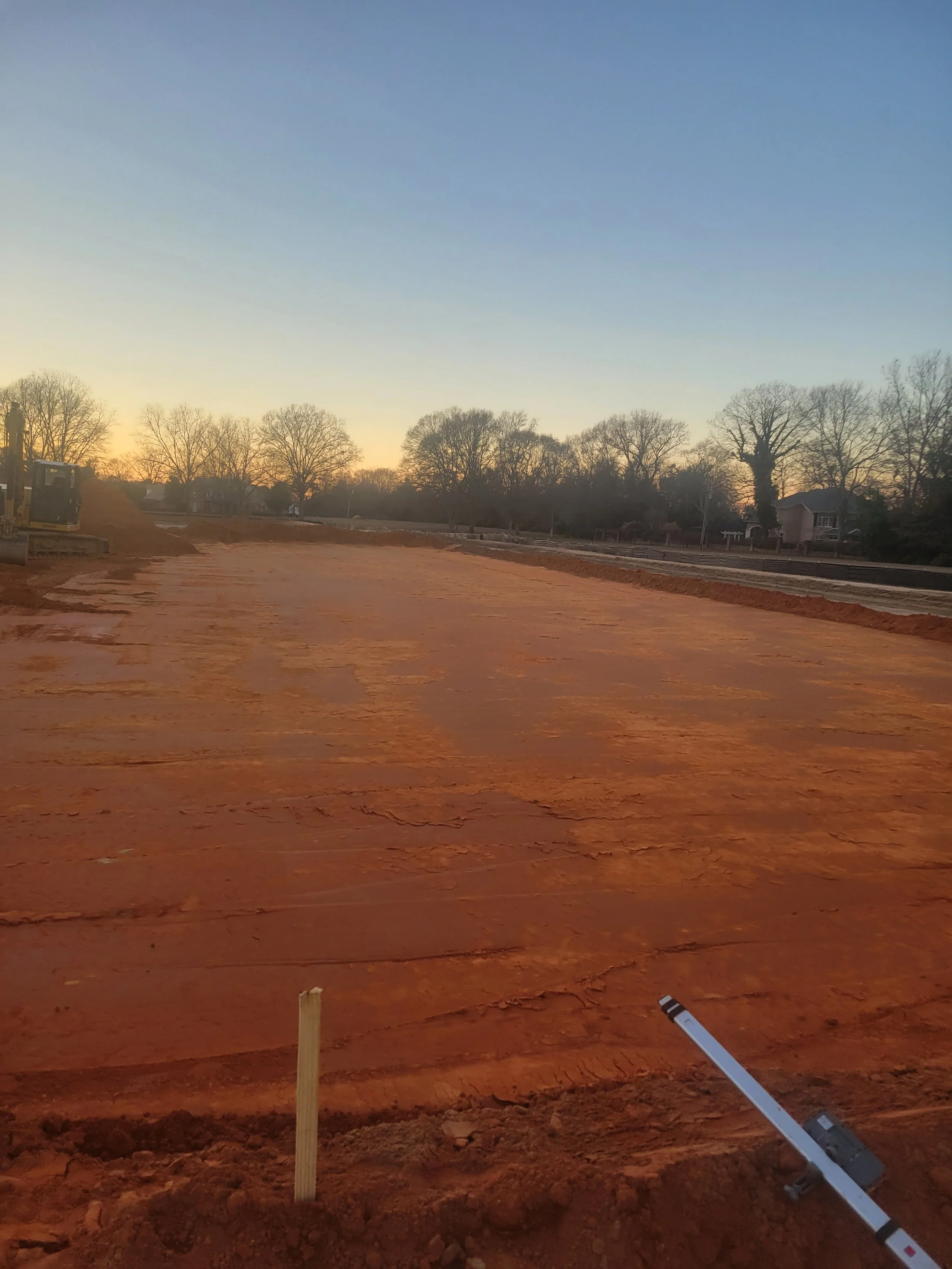 A construction site during sunset with a cleared, flat dirt surface, some construction equipment on the left, and trees and houses in the background.