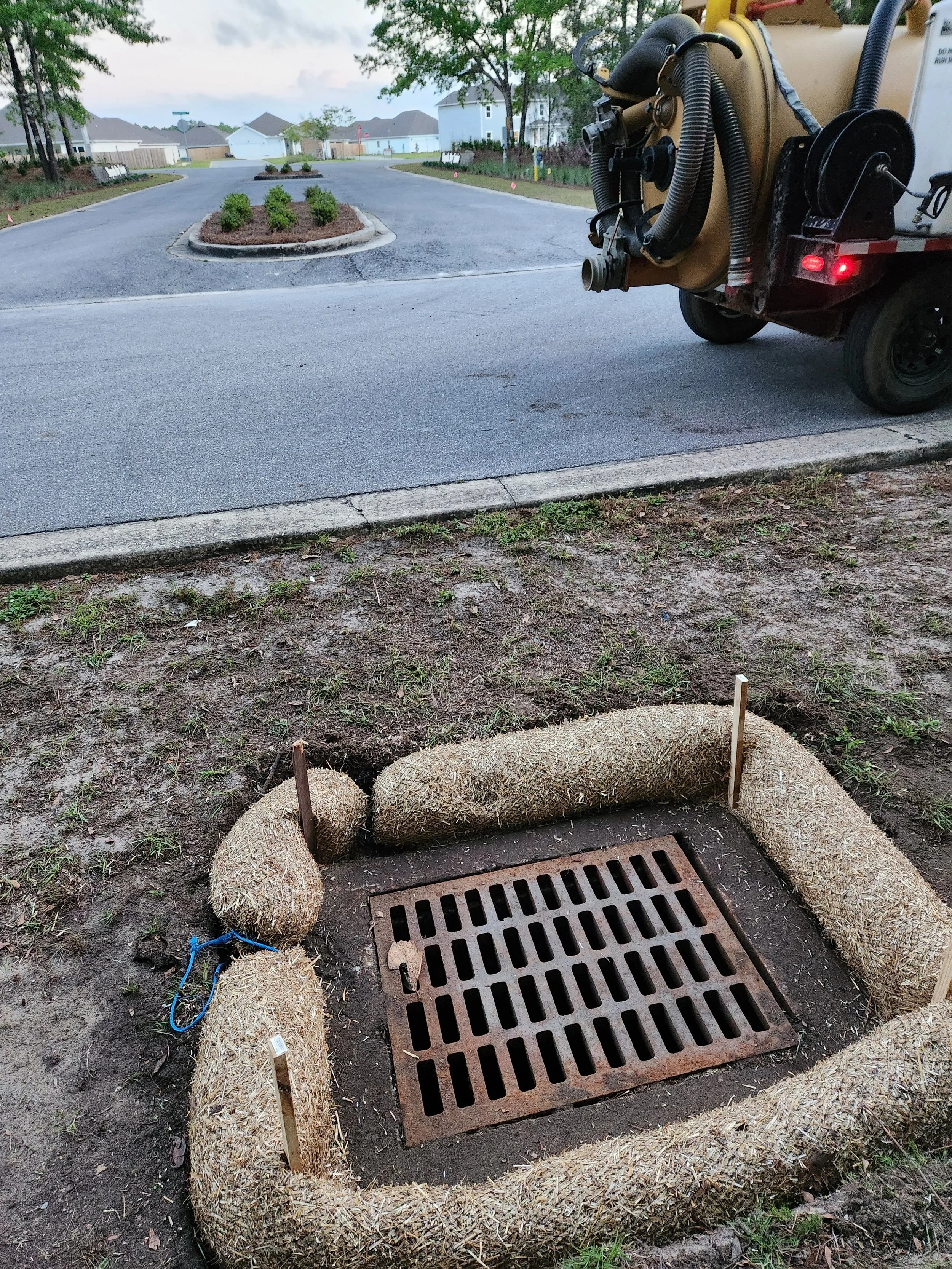 Freshly installed school bus stop cover with edging made of hay bales around a storm drain, stick markers, and a construction string tied to the hay bales.