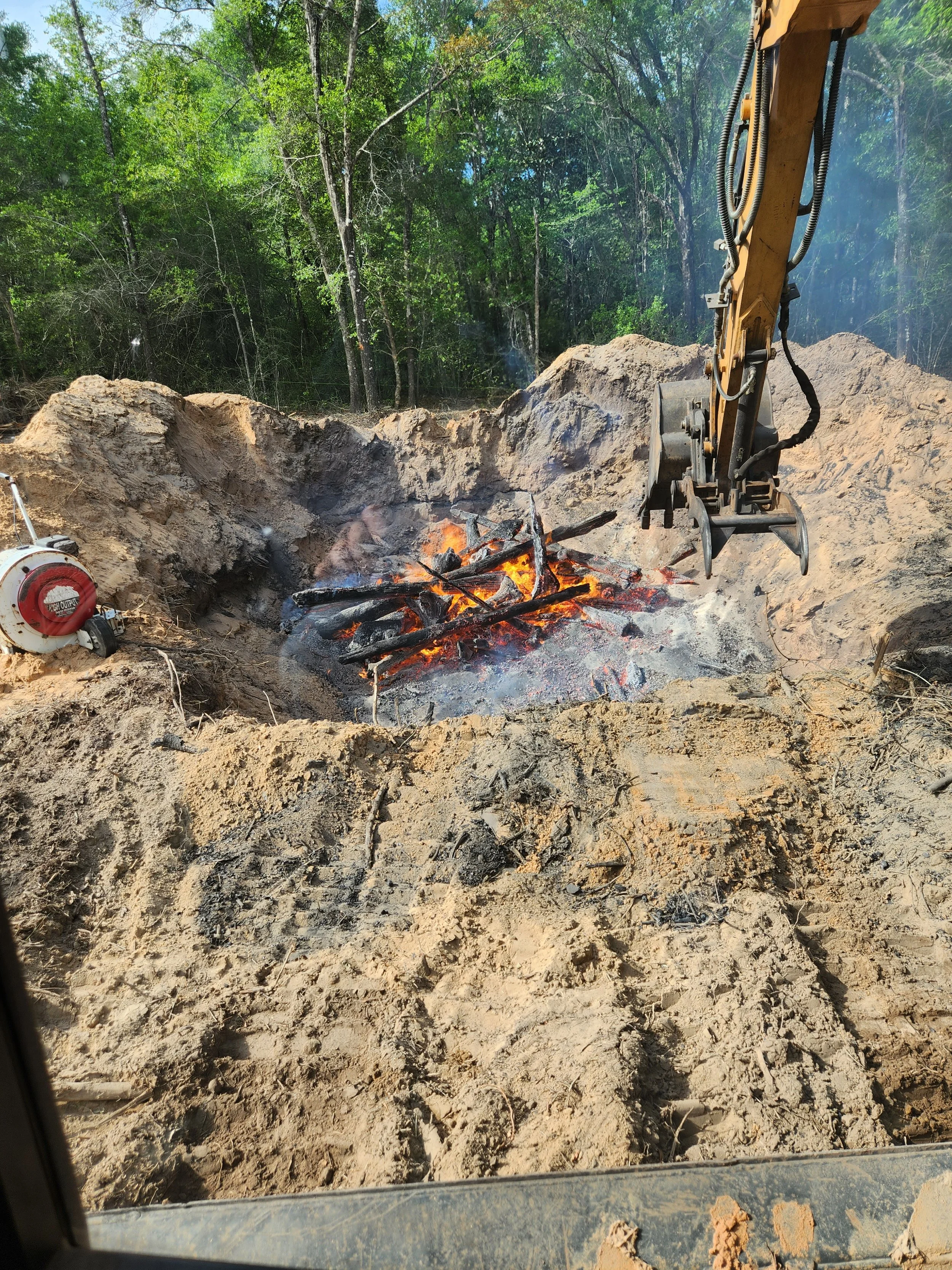 Excavator burning a pile of wood logs in a pit at a construction site surrounded by green trees.
