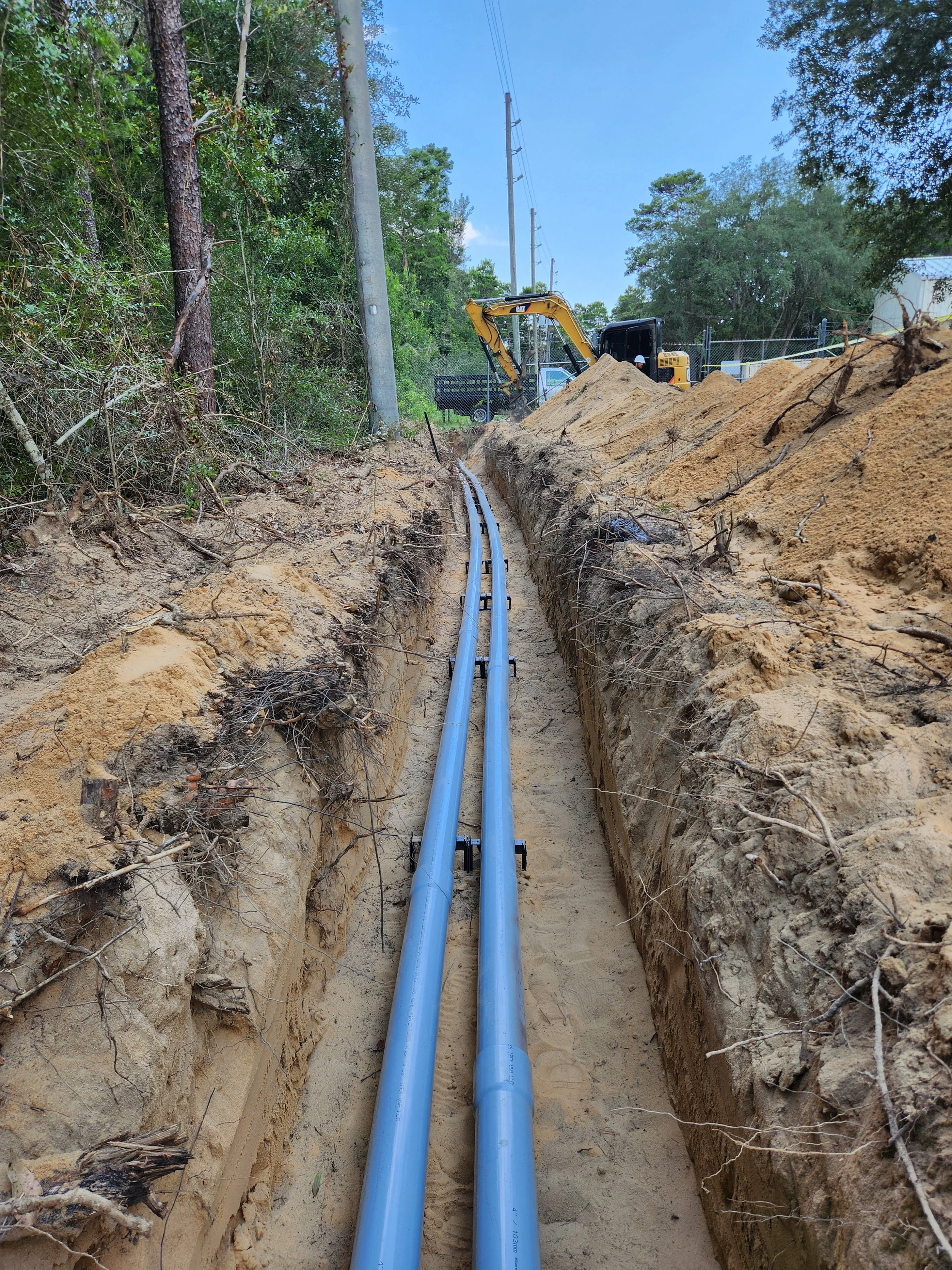 Construction site with two blue pipes laid in a trench, a small excavator in the background, along with trees, utility poles, and a blue sky.