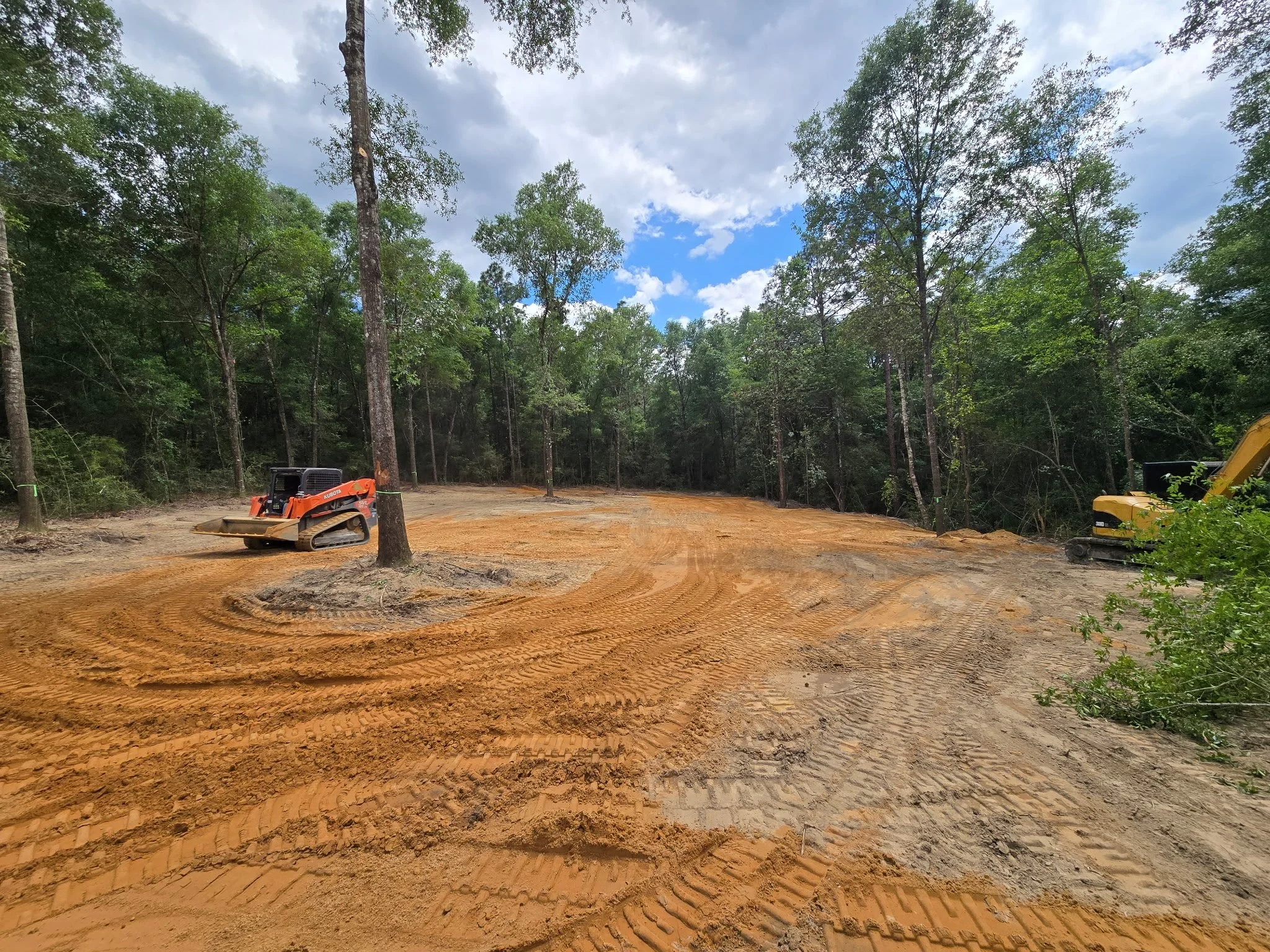 Construction site in a forest with visible soil and two excavators, one orange and one yellow, working on clearing the land under a partly cloudy sky.