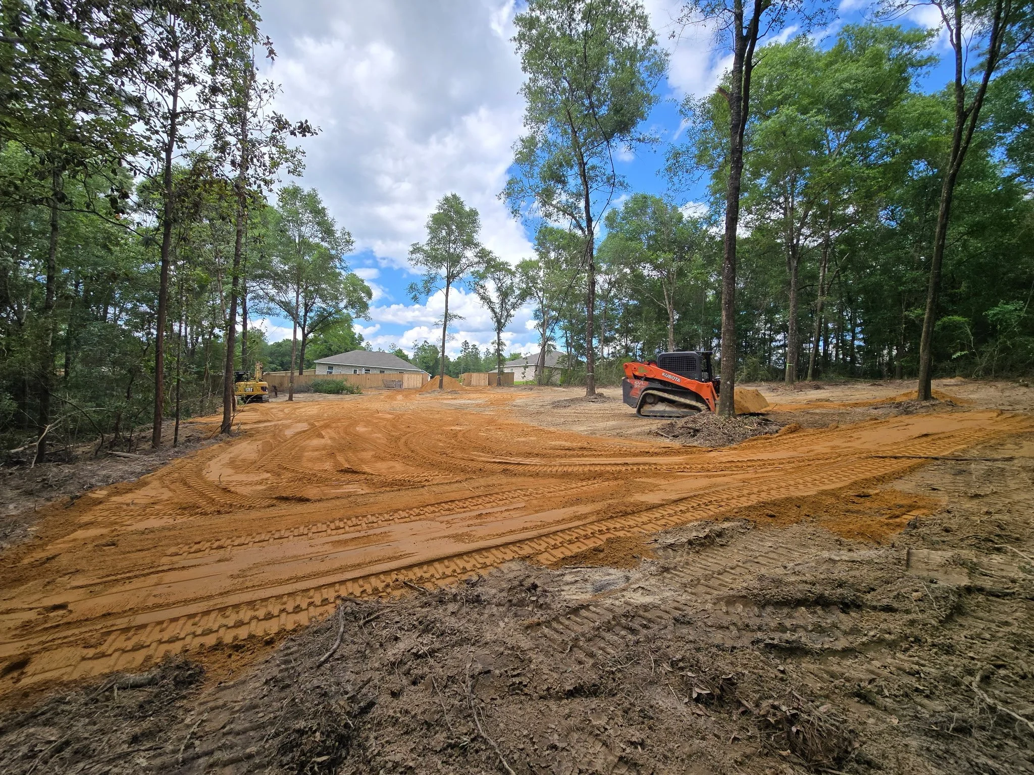 A construction site with orange dirt, tire tracks, and a small orange bulldozer. Trees and houses are in the background under a partly cloudy sky.