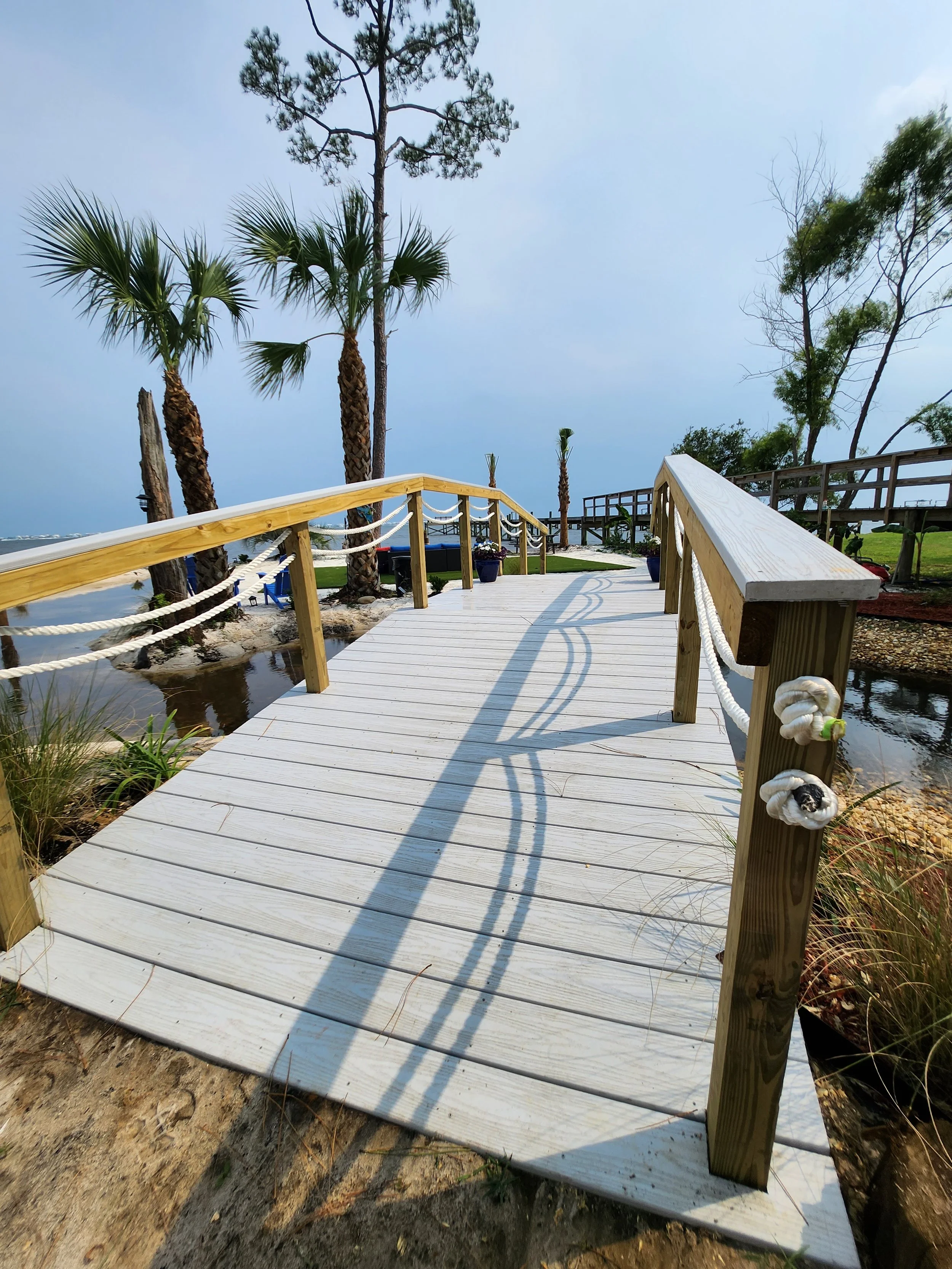 Wooden dock with handrails on either side extending over a pond or small lake, with tall palm trees and other trees in the background on a cloudy day.