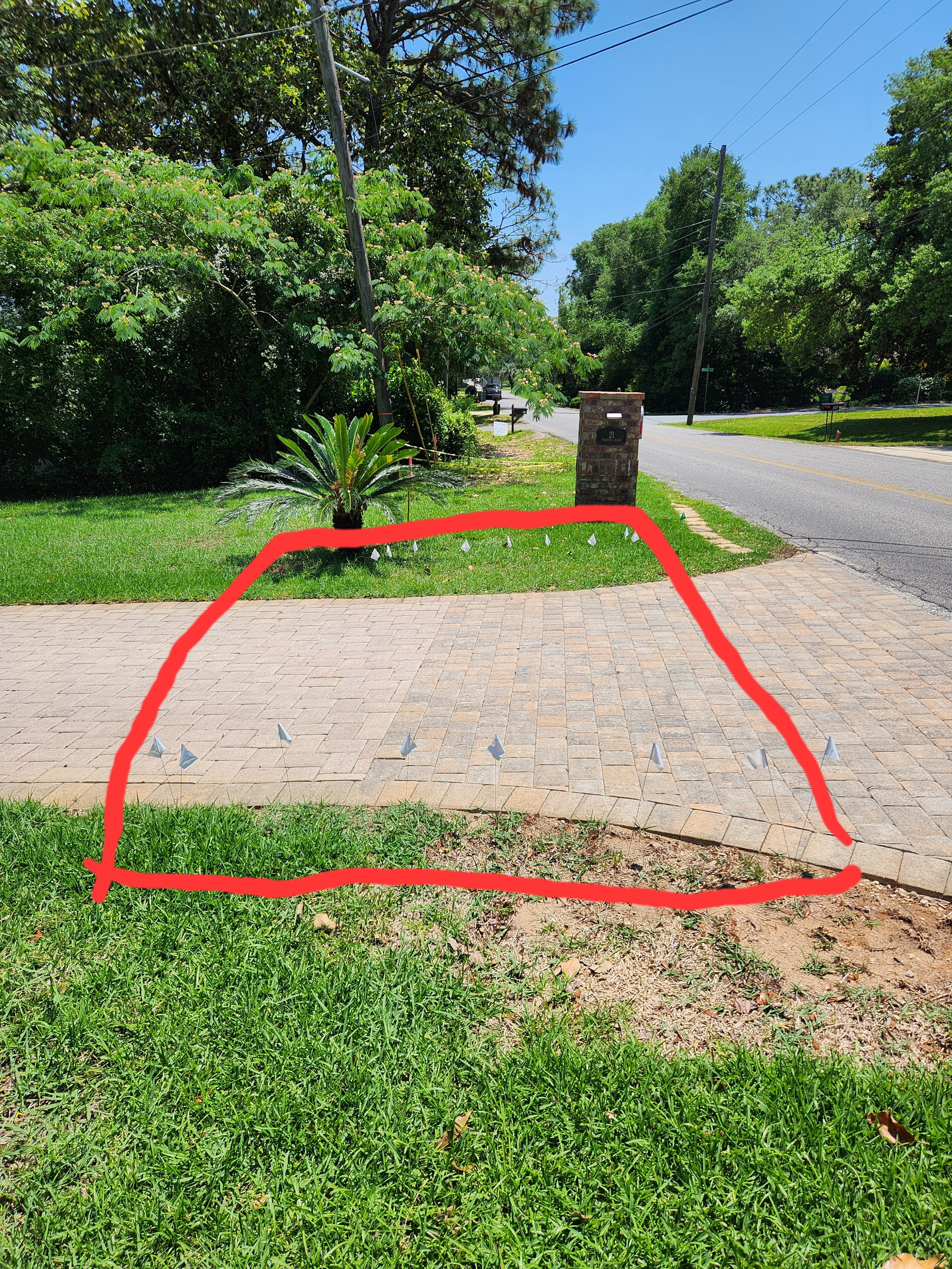 A suburban driveway bordered by a small patch of grass and a brick sidewalk. The sidewalk has small, white, cone-shaped markings at regular intervals. To the right, a street with a black asphalt surface and yellow dividing lines, with a brick mailbox