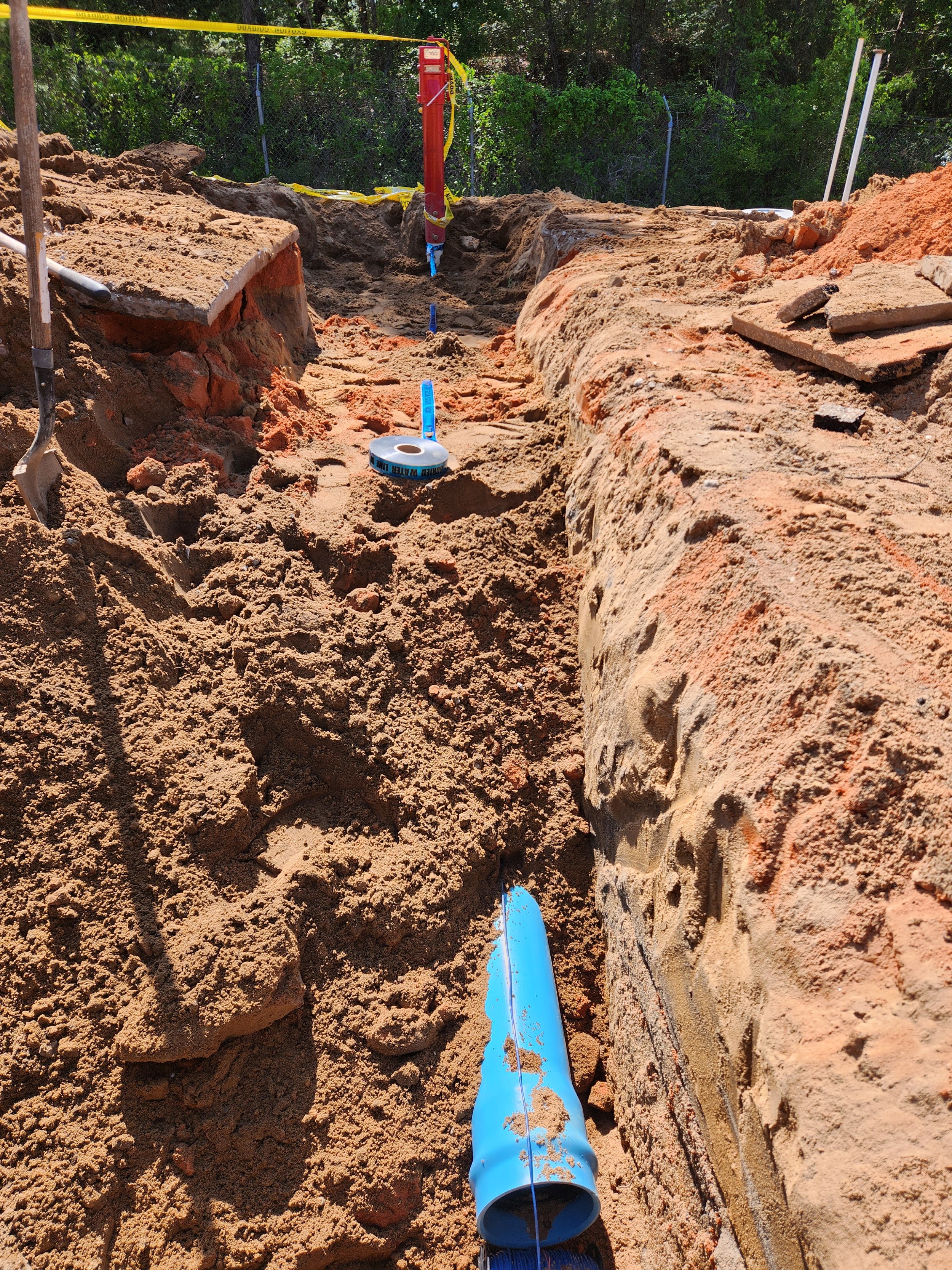 Construction site with excavated trench, blue pipe, and surveying tools, surrounded by dirt and some greenery in the background.