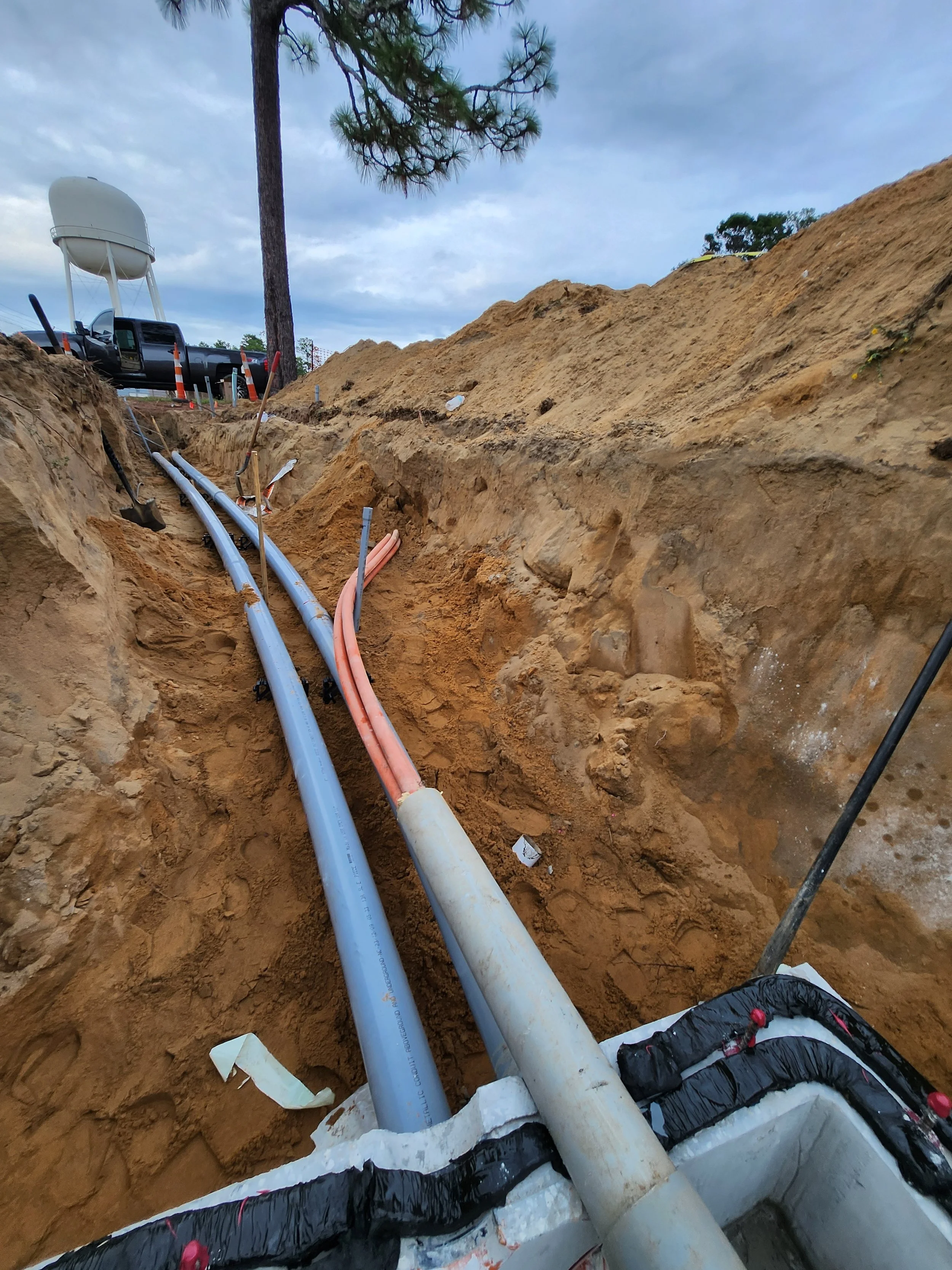Underground utility pipes, including blue and orange conduits, laid in a trench at a construction site, with a pickup truck and weathered sky in the background.