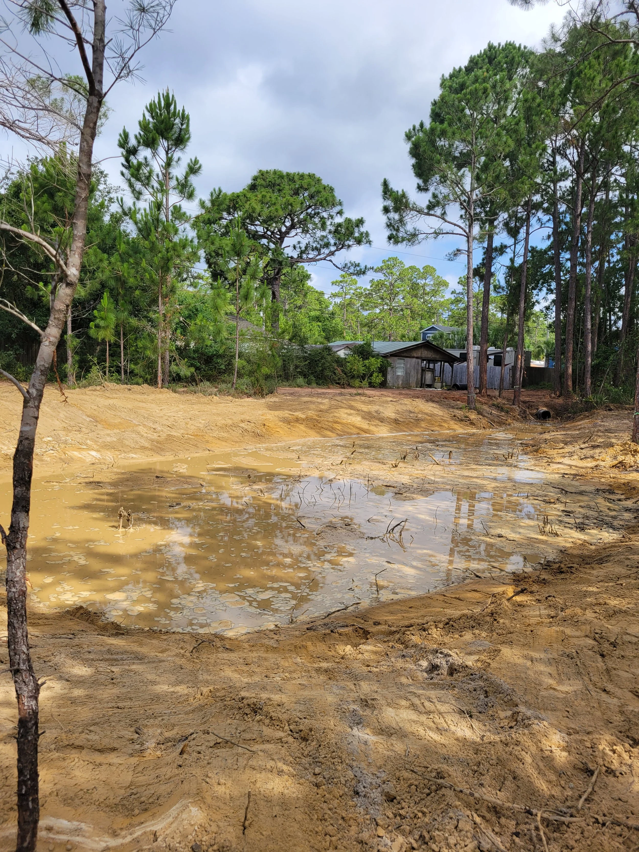 A muddy, water-filled excavation site with green trees and a small wooden building in the background under a cloudy sky.
