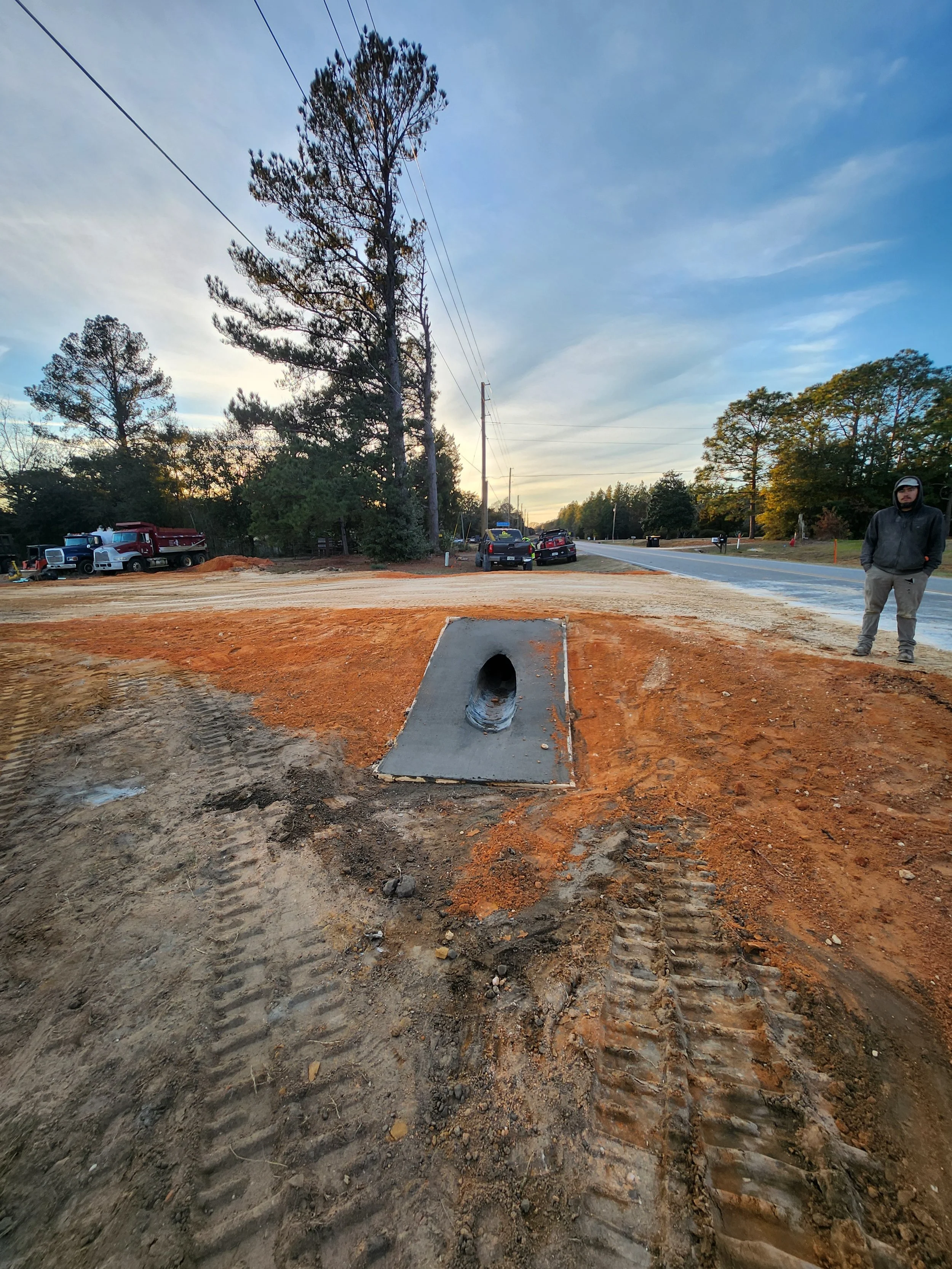Construction site with a concrete drainage pipe embedded in the ground, orange dirt and tire tracks in the foreground, parked trucks and cars, trees, power lines, and a person standing on the right side wearing a hoodie and cap.