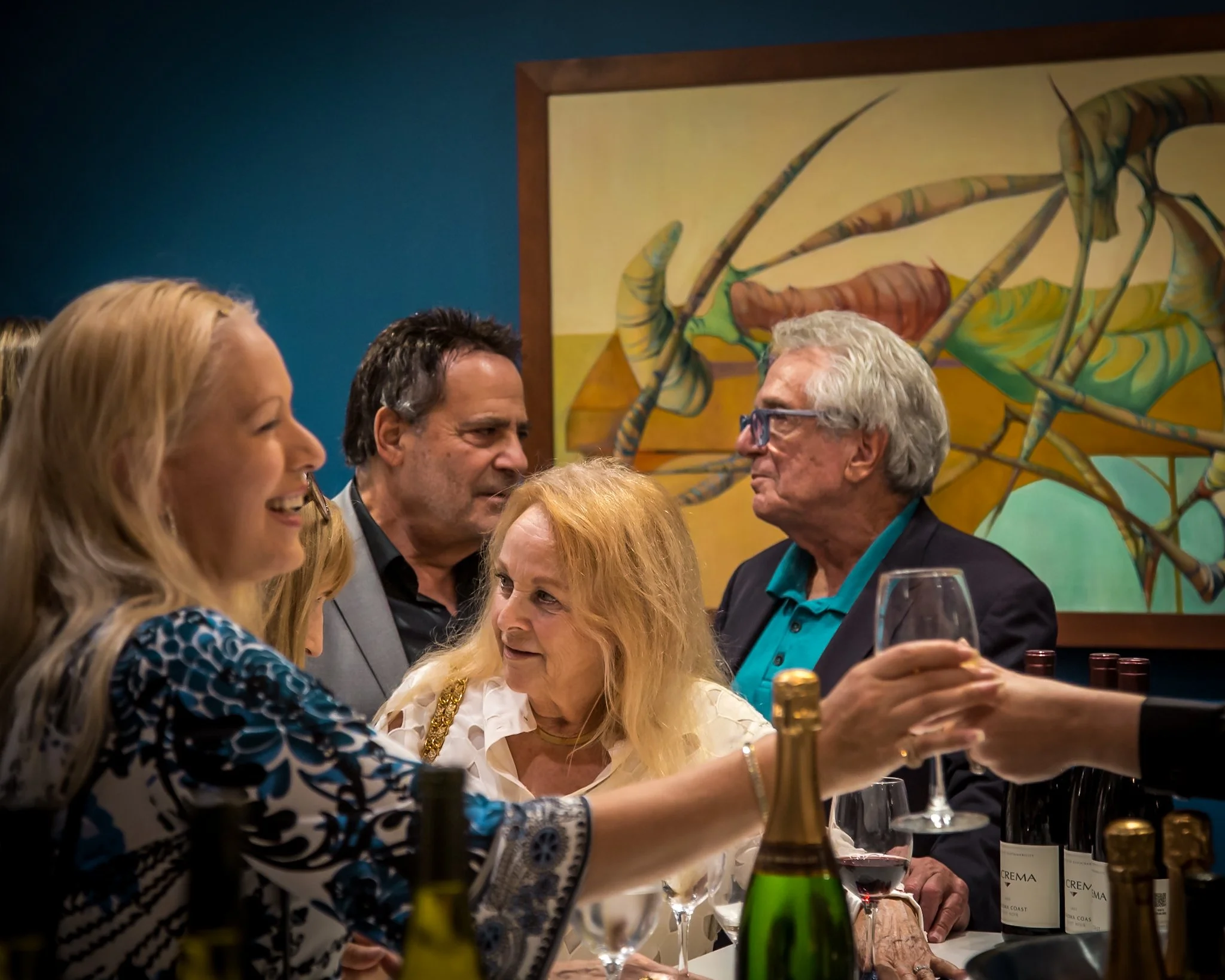 People gathered around a table at a social event, exchanging drinks and engaging in conversation, with a colorful abstract painting in the background.
