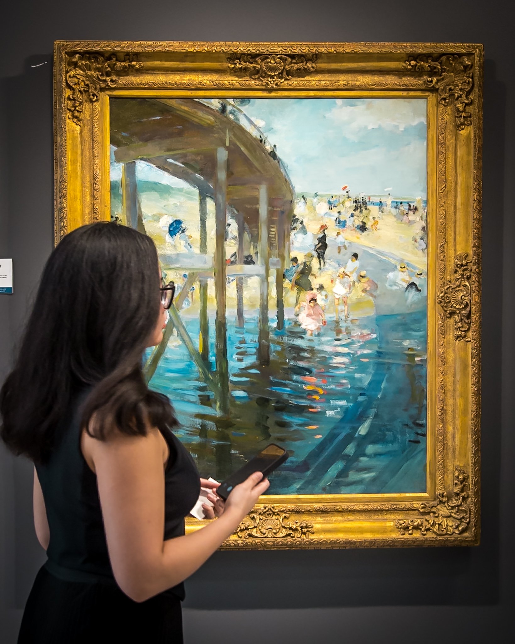 Woman looking at a painting of a beach scene with a pier, people, and water, in a gold ornate frame.
