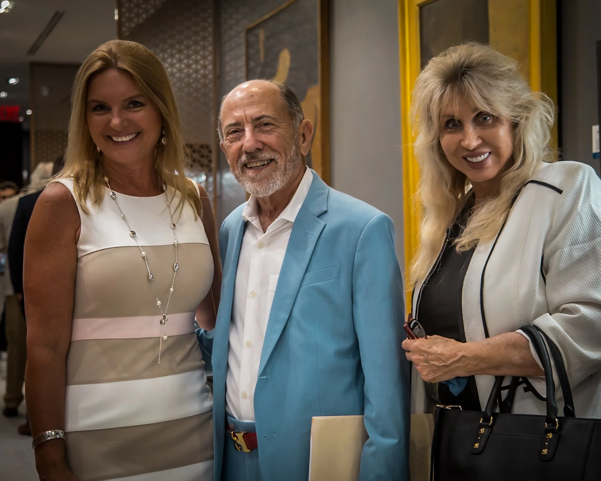 Three well-dressed people smiling at an indoor event, with two women and one man in the center, all facing the camera.