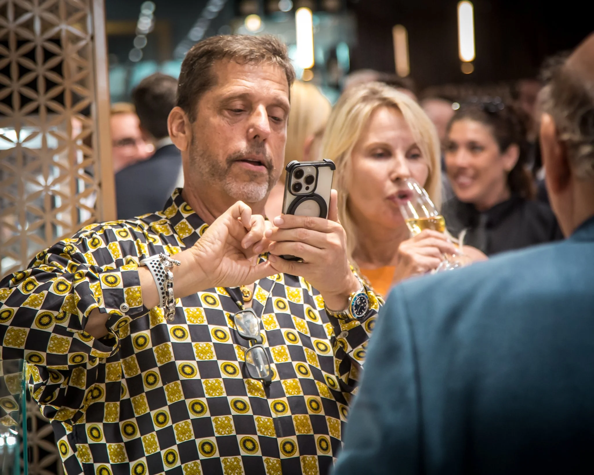 A man in a patterned shirt is taking a photo with his phone at a social gathering. A woman next to him is holding a glass of champagne, and other guests are visible in the background.