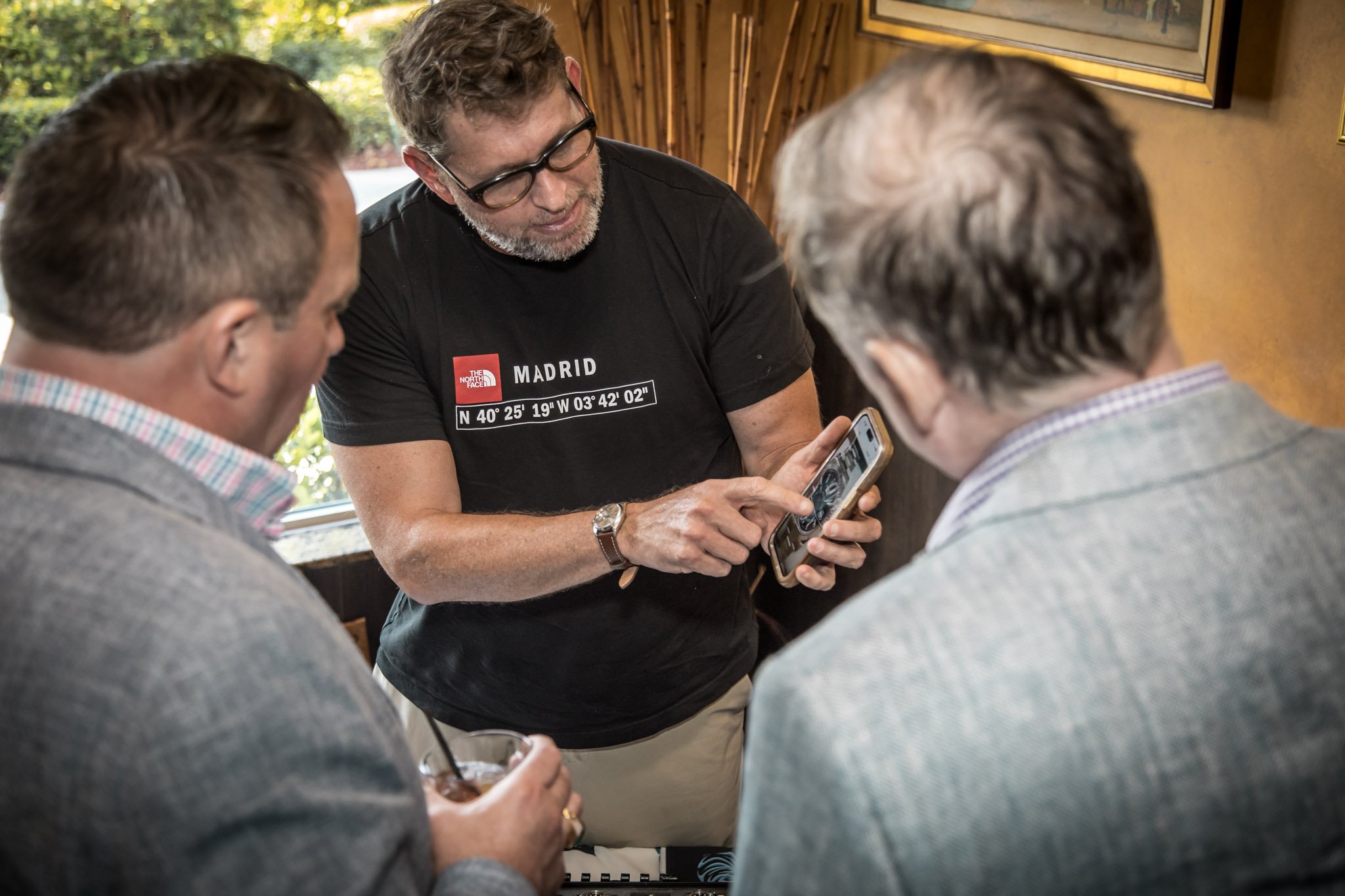 Three men gathered around a smartphone, one of them showing something on the screen to the other two inside a room with wooden walls and framed pictures.
